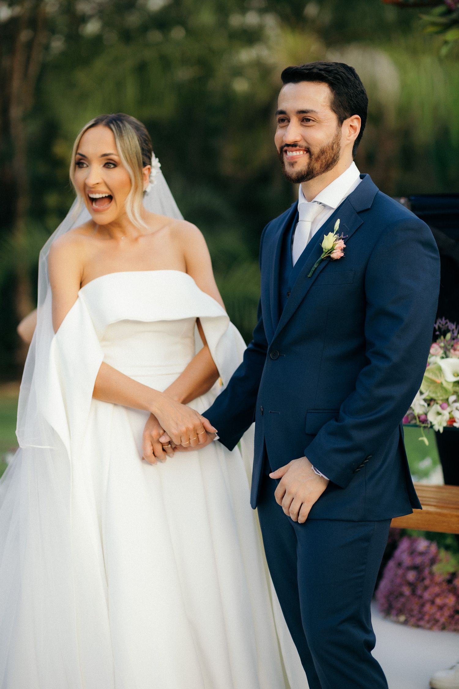 Joyful bride and groom holding hands and laughing during outdoor wedding ceremony.