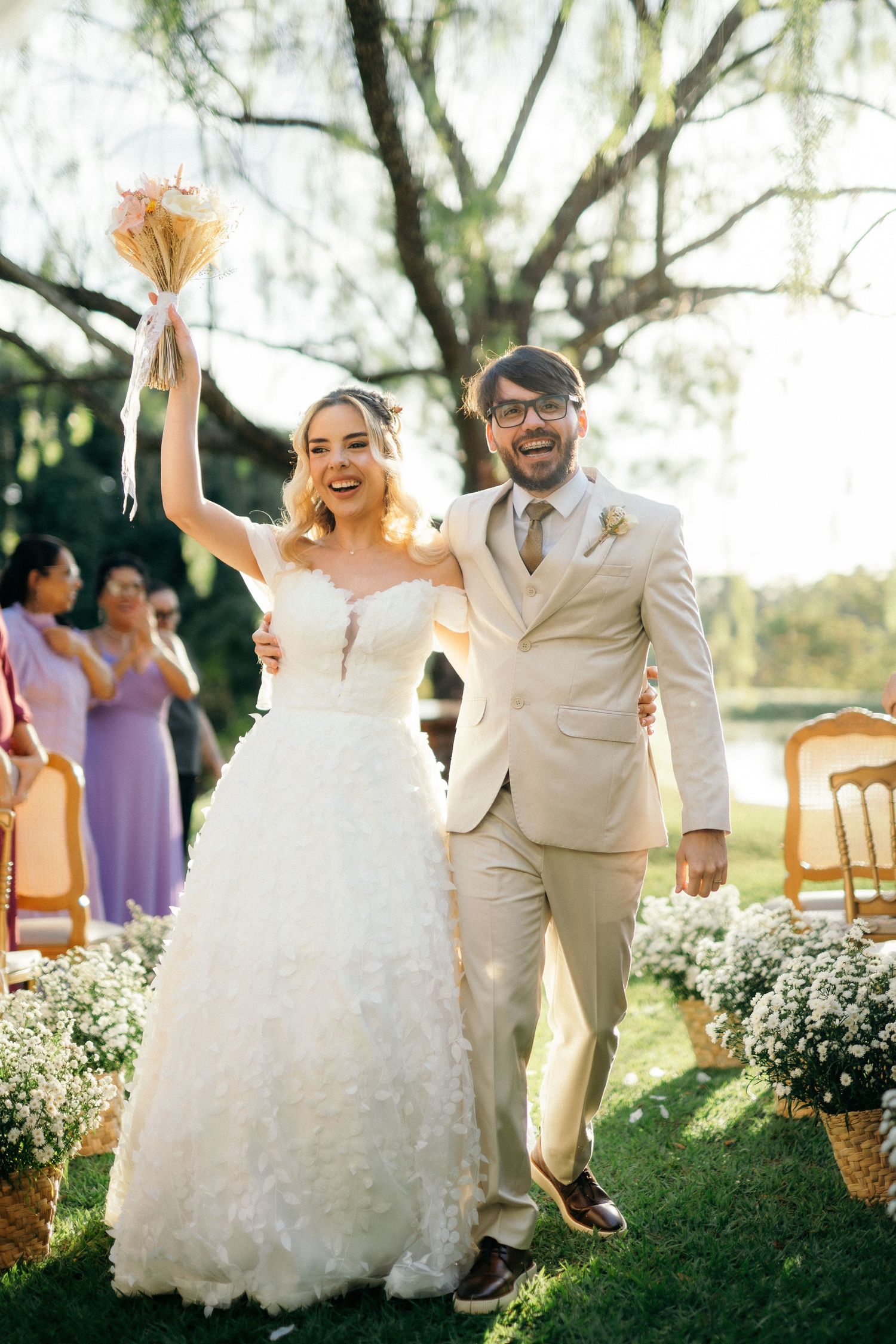 Joyful bride in white gown raises bouquet beside smiling groom in beige suit during outdoor wedding ceremony.