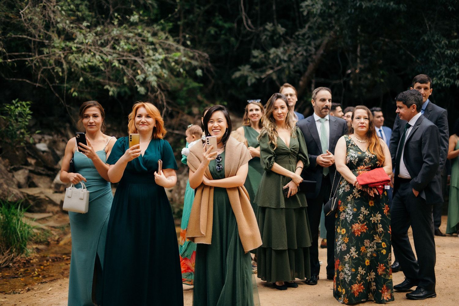 Group of wedding guests in green dresses applauding outdoors near forest waterfall setting.