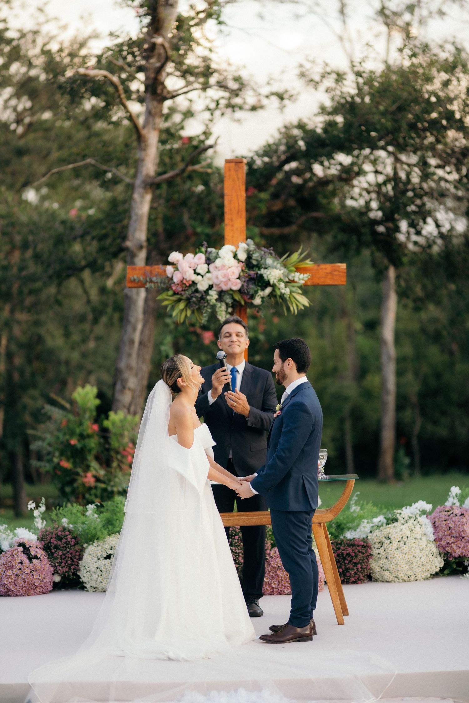 Bride and groom exchange vows before wooden cross with flowers at outdoor wedding ceremony.