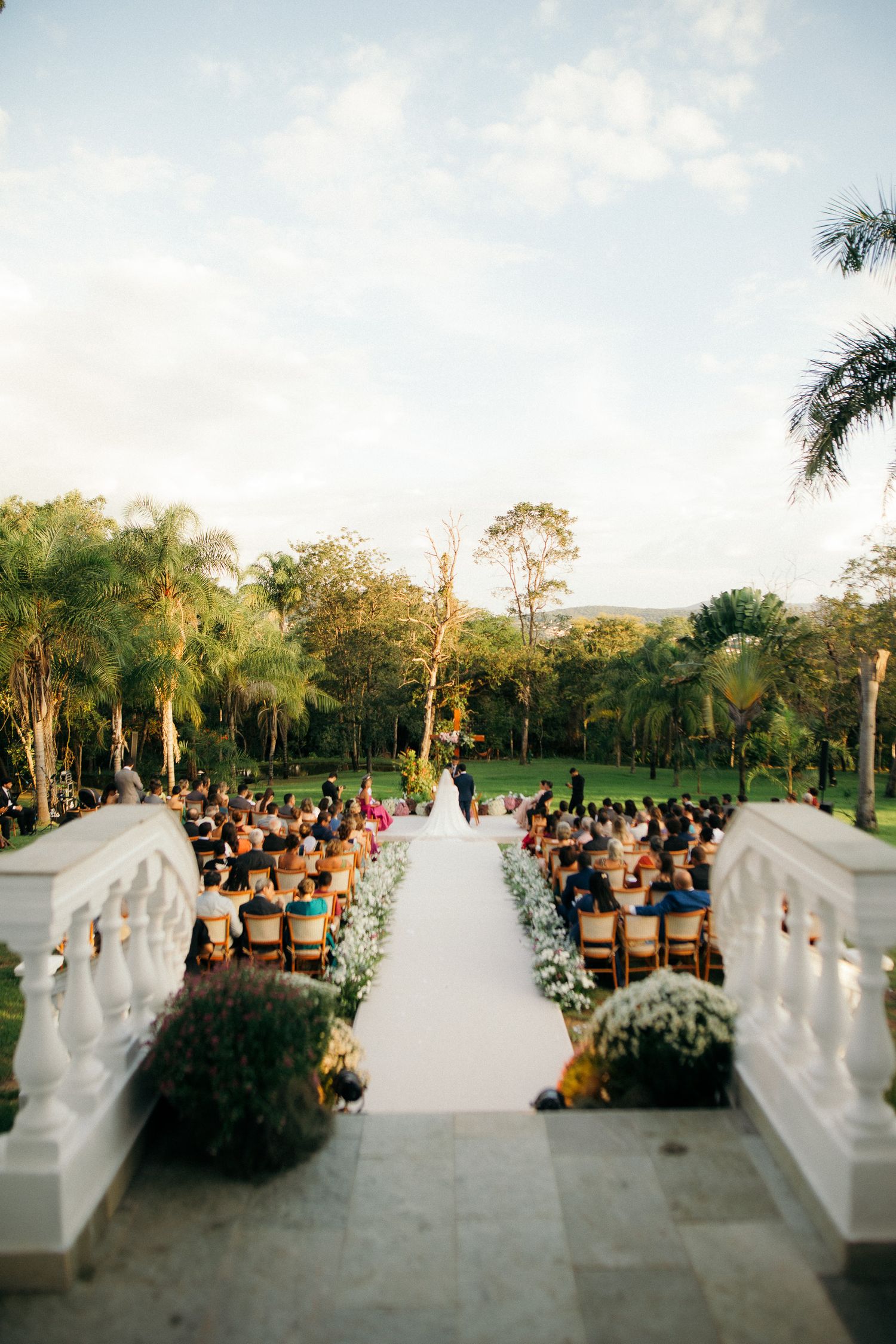Aerial view of an outdoor wedding ceremony with white aisle, seated guests, and palm trees at golden hour.