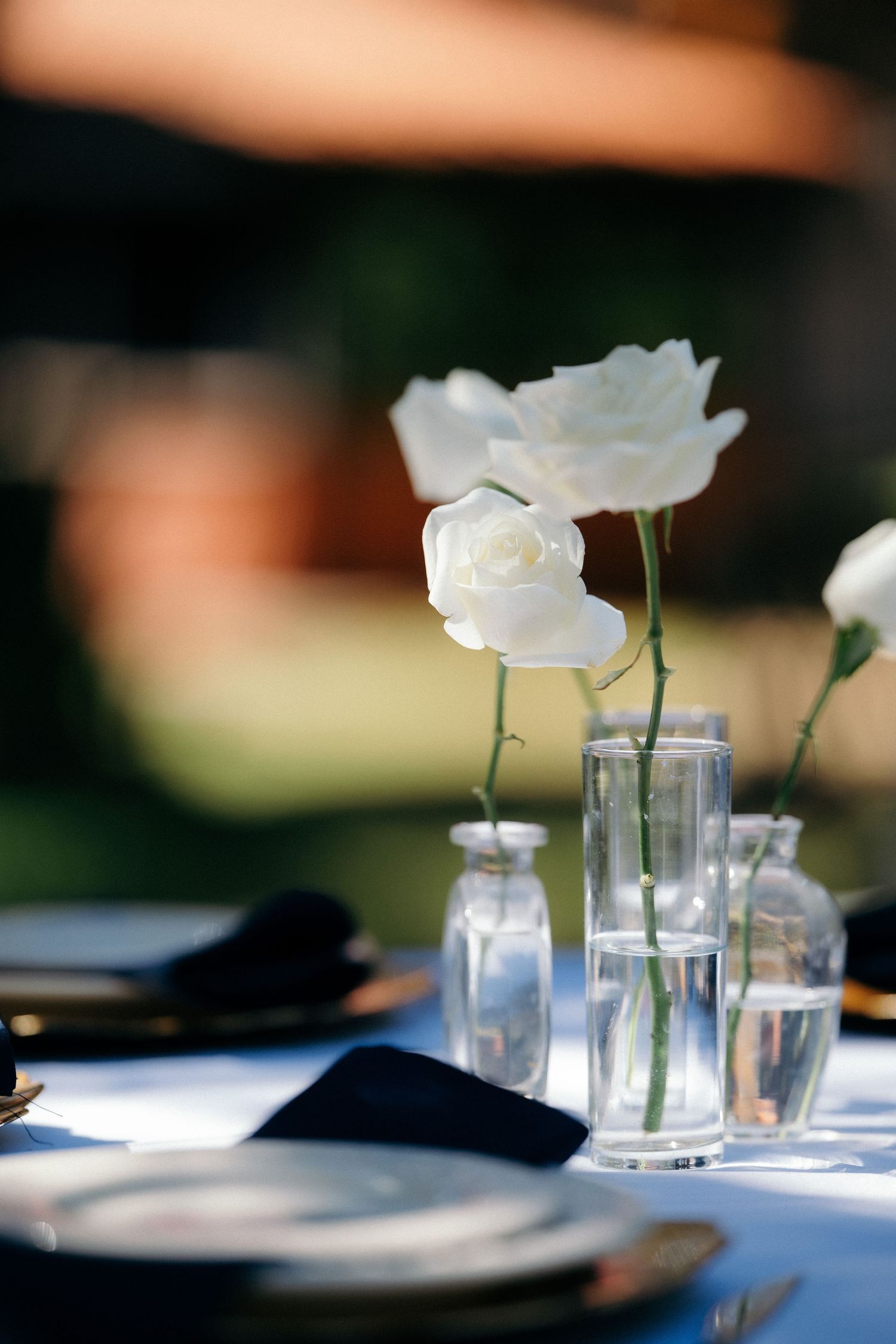 White roses in small glass vases on an elegant outdoor wedding table setting with dark napkins.