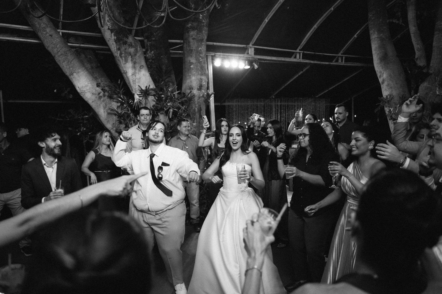 Black and white photo of bride and groom dancing joyfully with guests at an outdoor wedding reception.