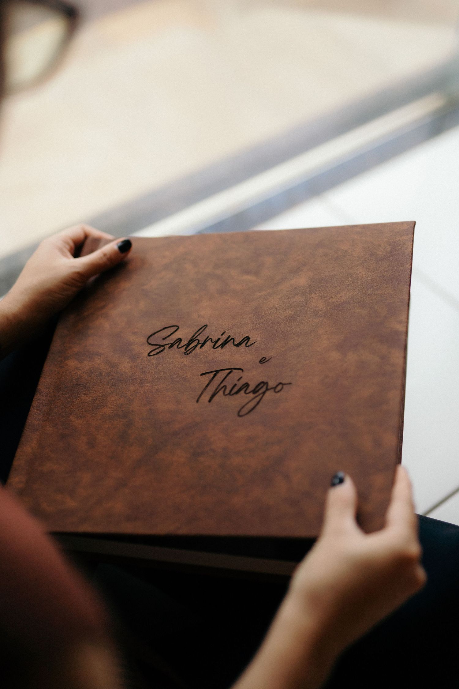 Hands holding a brown leather wedding photo album engraved with the names Sabrina and Thiago.
