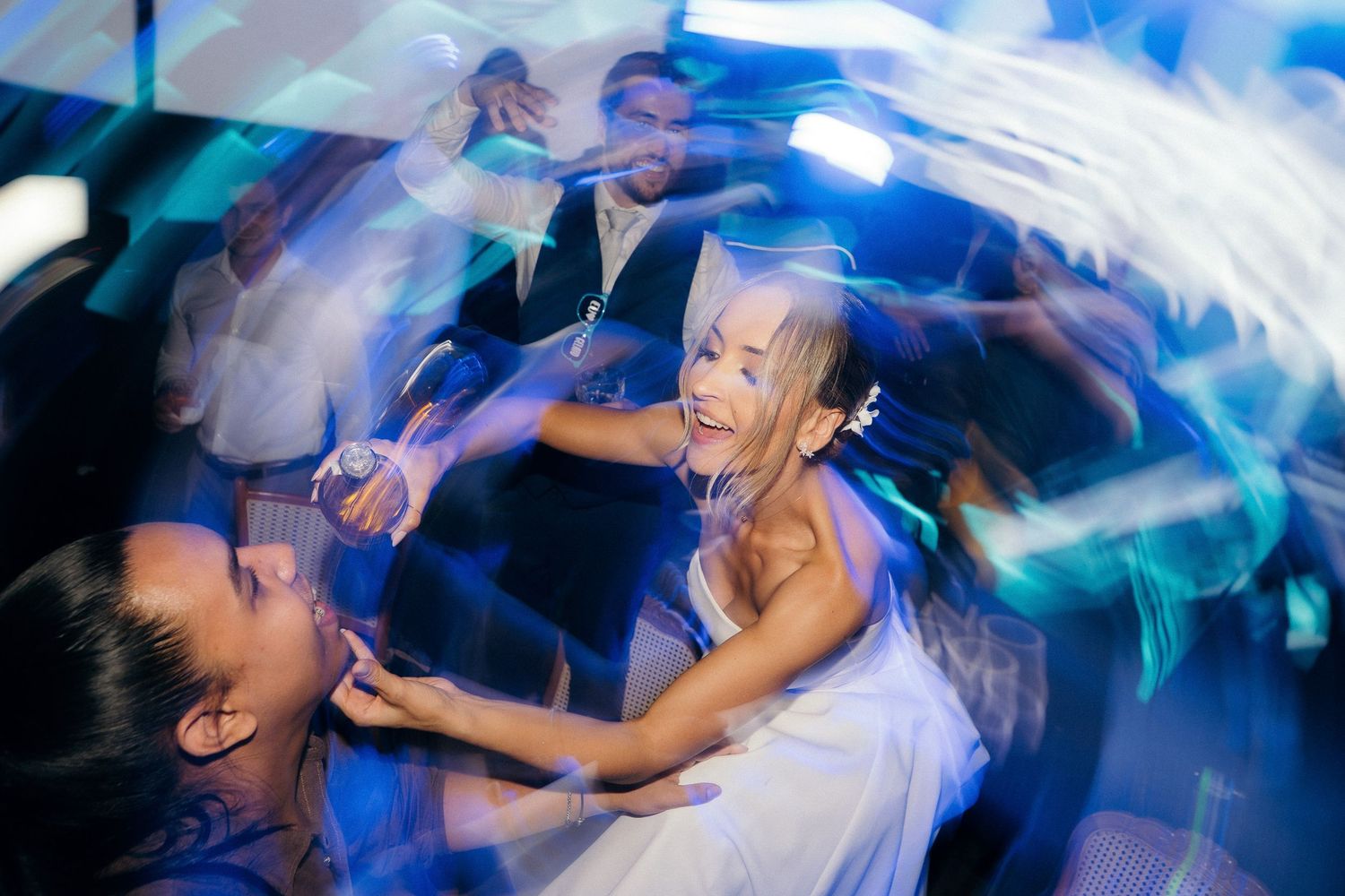 Bride in white gown dances joyfully with guests under colorful lights at lively wedding reception.