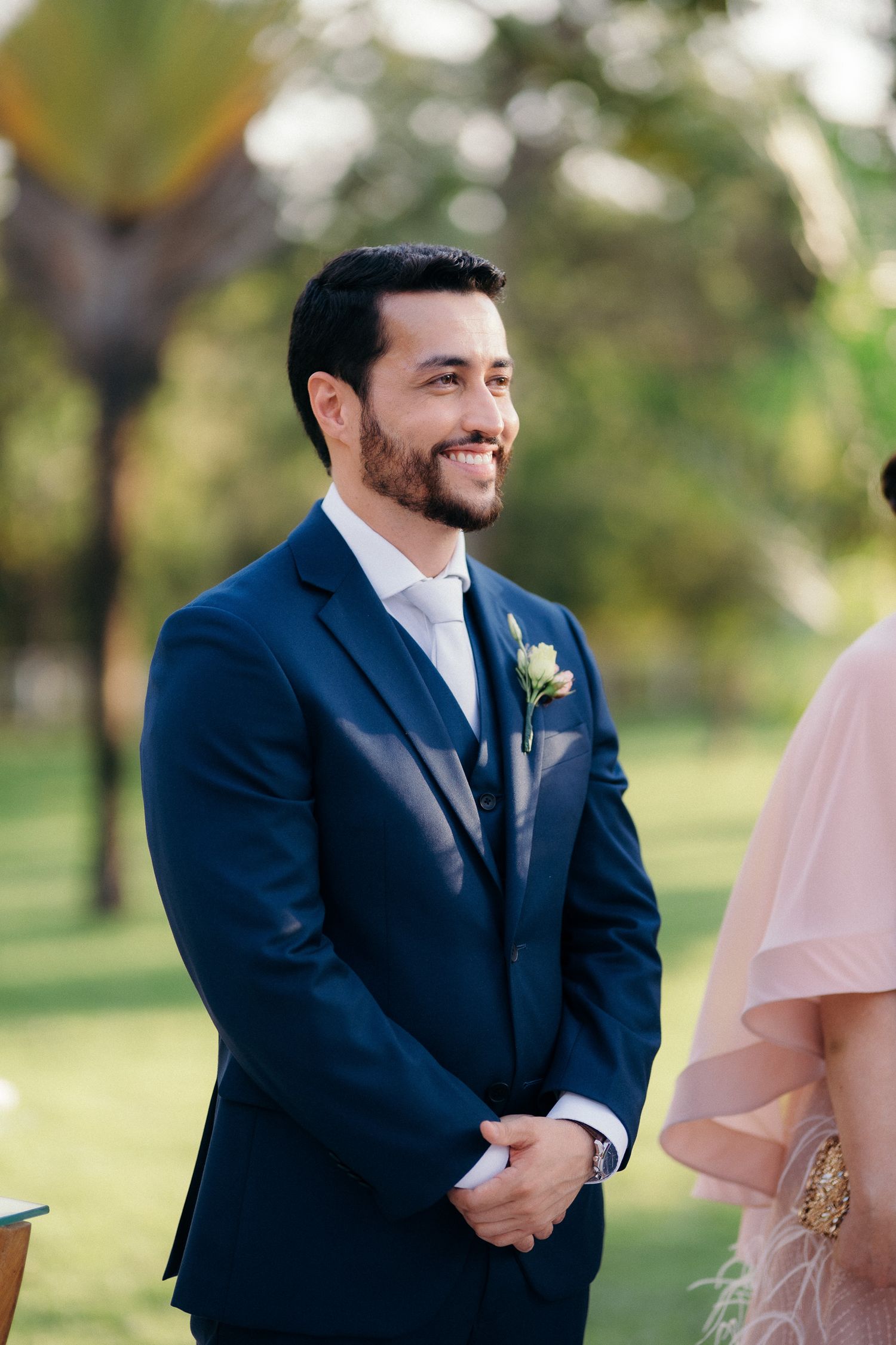 Smiling groom in navy suit with boutonniere watching bride walk down the aisle outdoors.