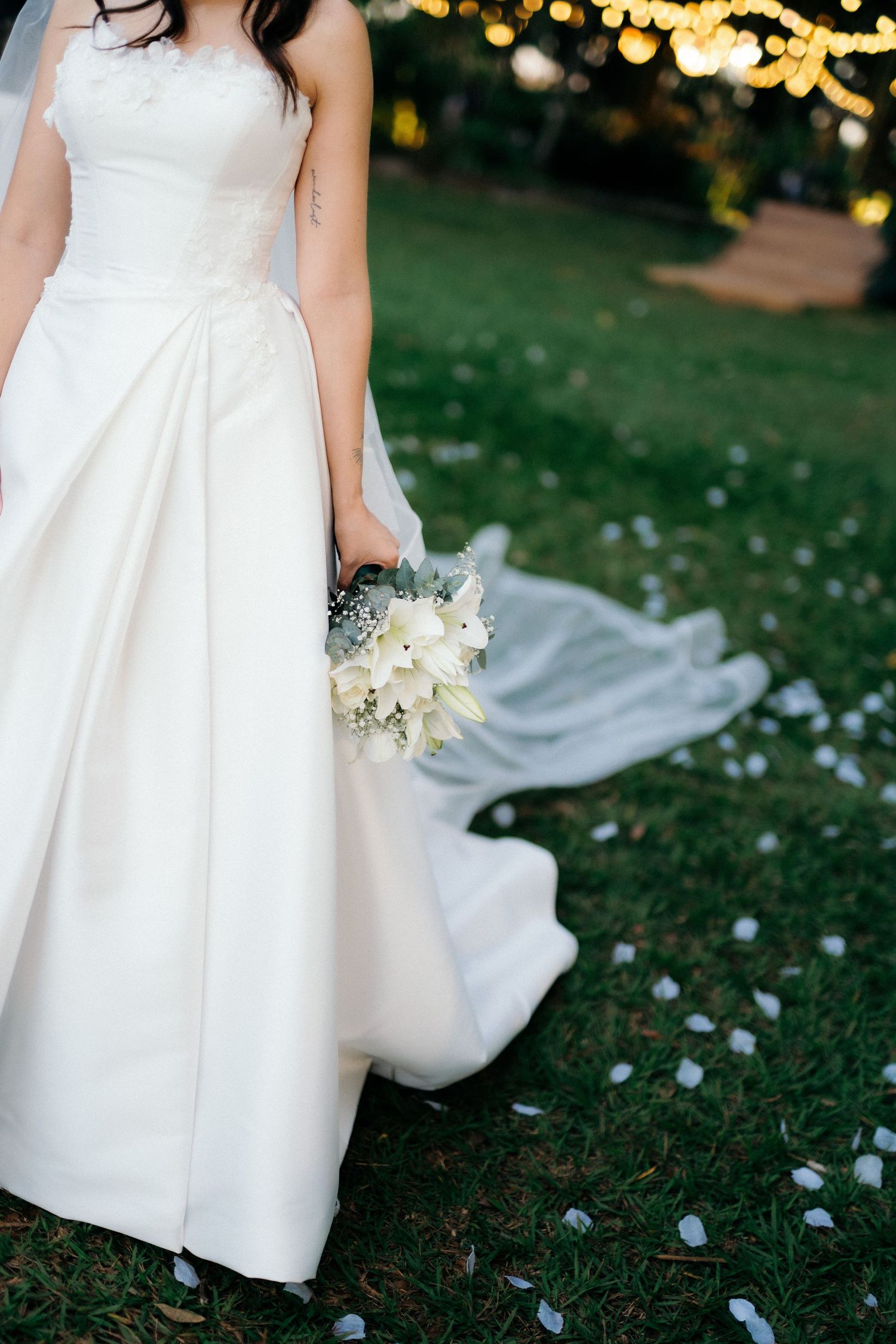 Bride in white ball gown holding white floral bouquet on grass with rose petals scattered.