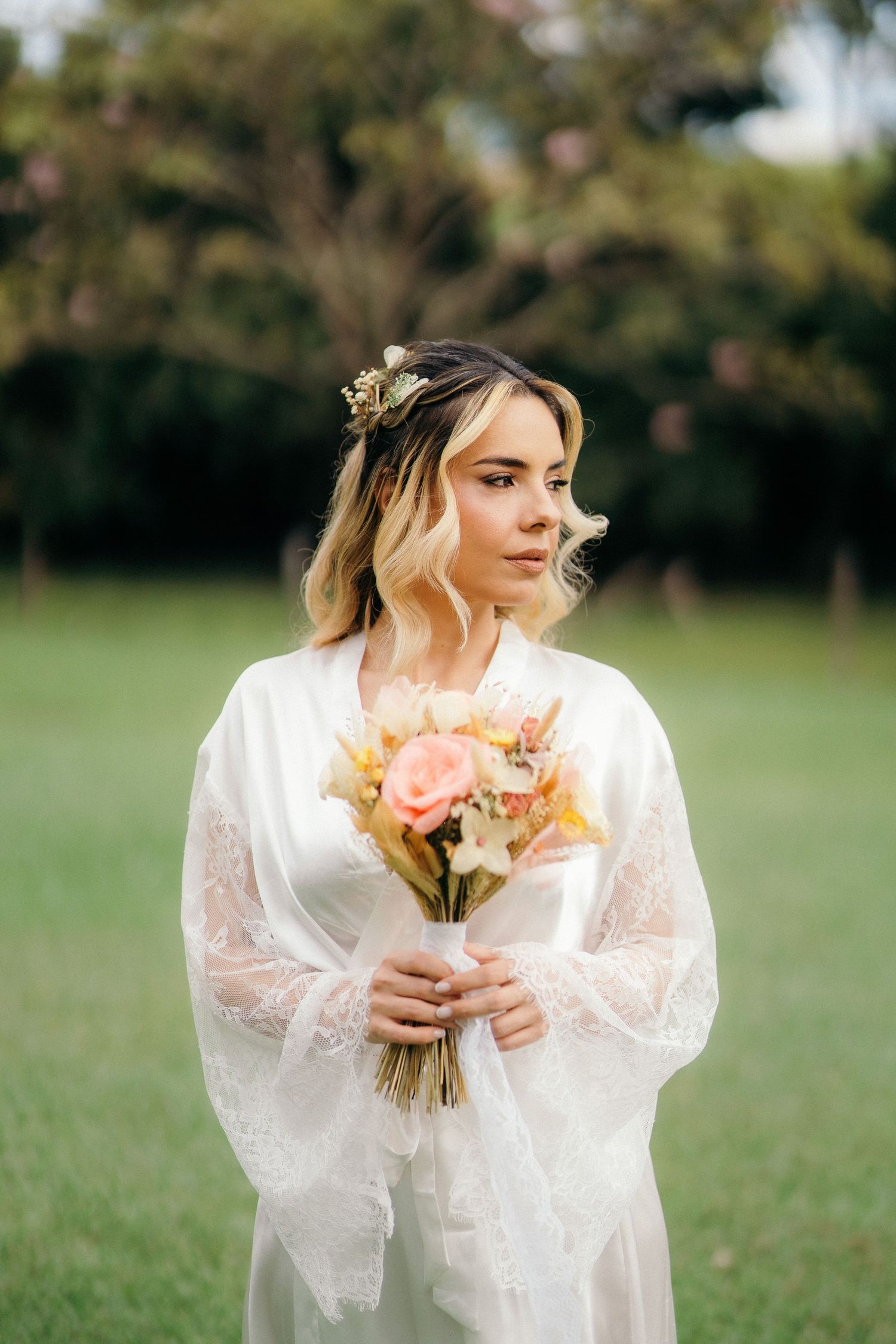 Bride in white lace robe holding a pink and peach floral bouquet with flowers in her hair, standing in a green garden.