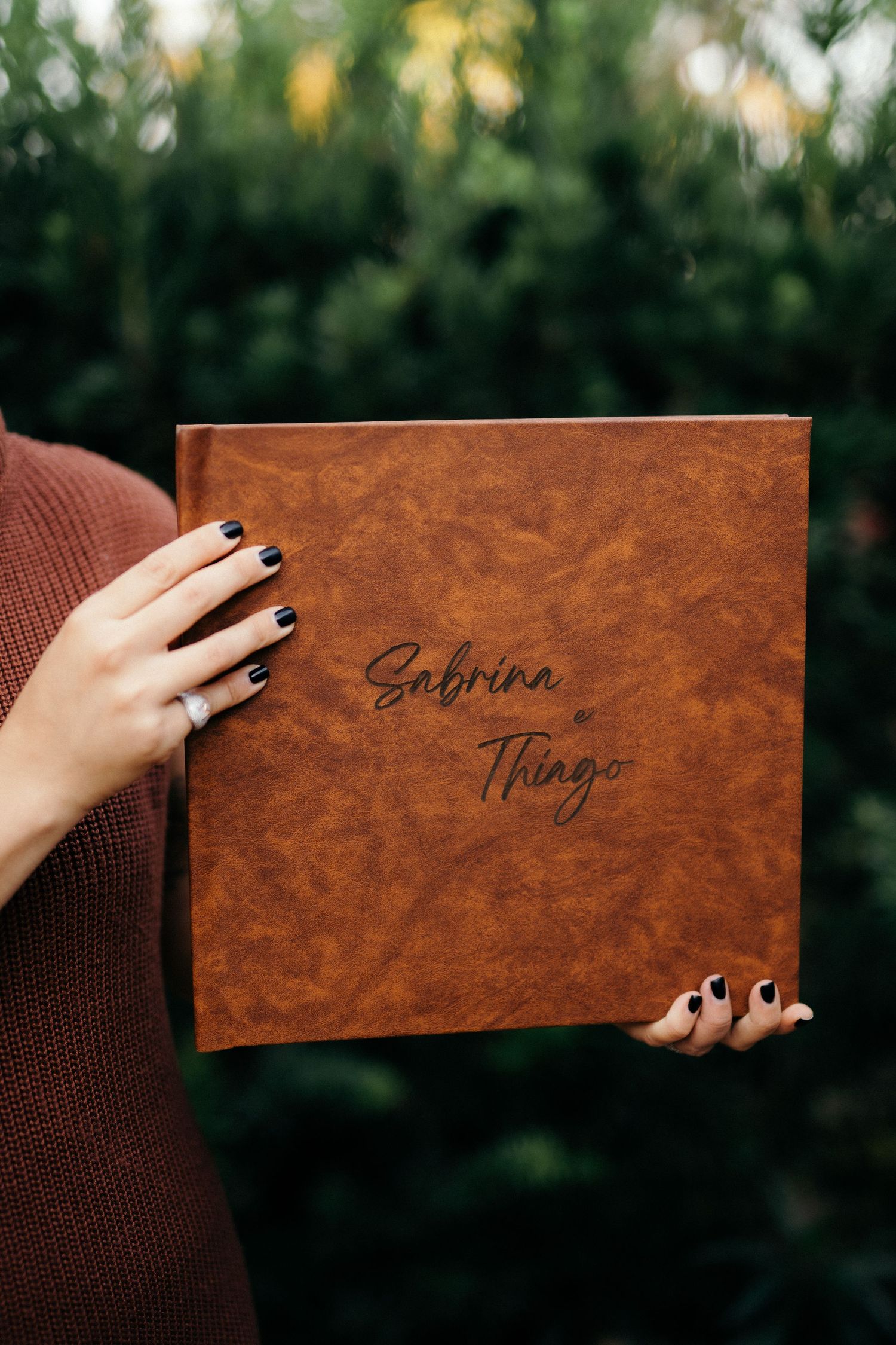 Hands with black nail polish holding a brown leather wedding album engraved with 