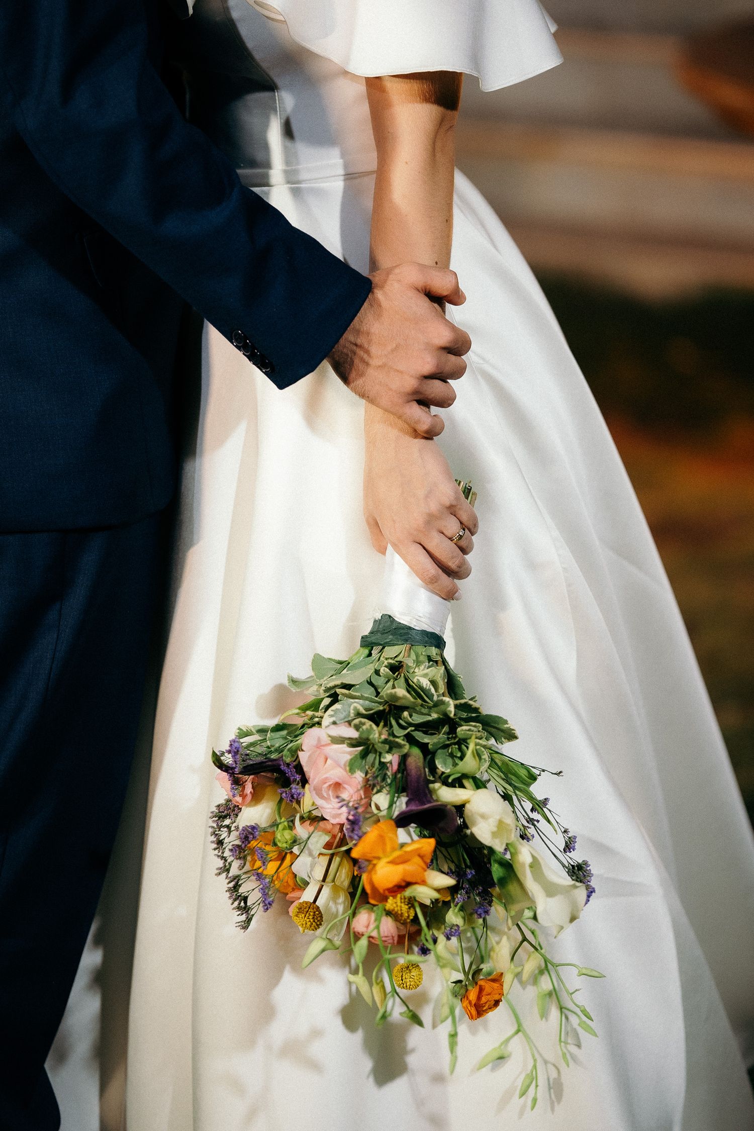 Bride and groom holding hands with a colorful wedding bouquet of wildflowers and greenery against a white bridal gown.
