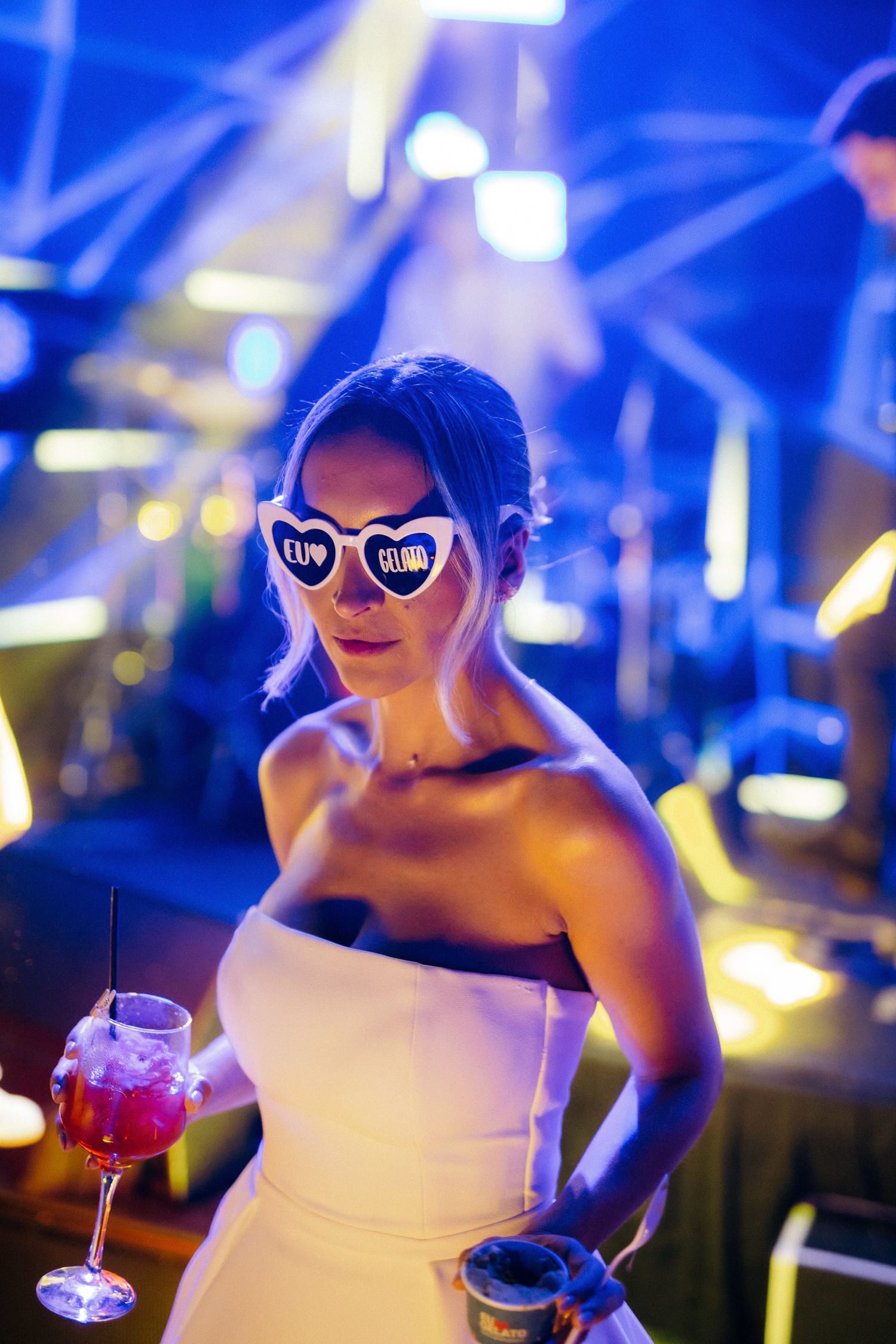 Bride in white strapless gown wears novelty heart sunglasses while holding cocktail at wedding reception.