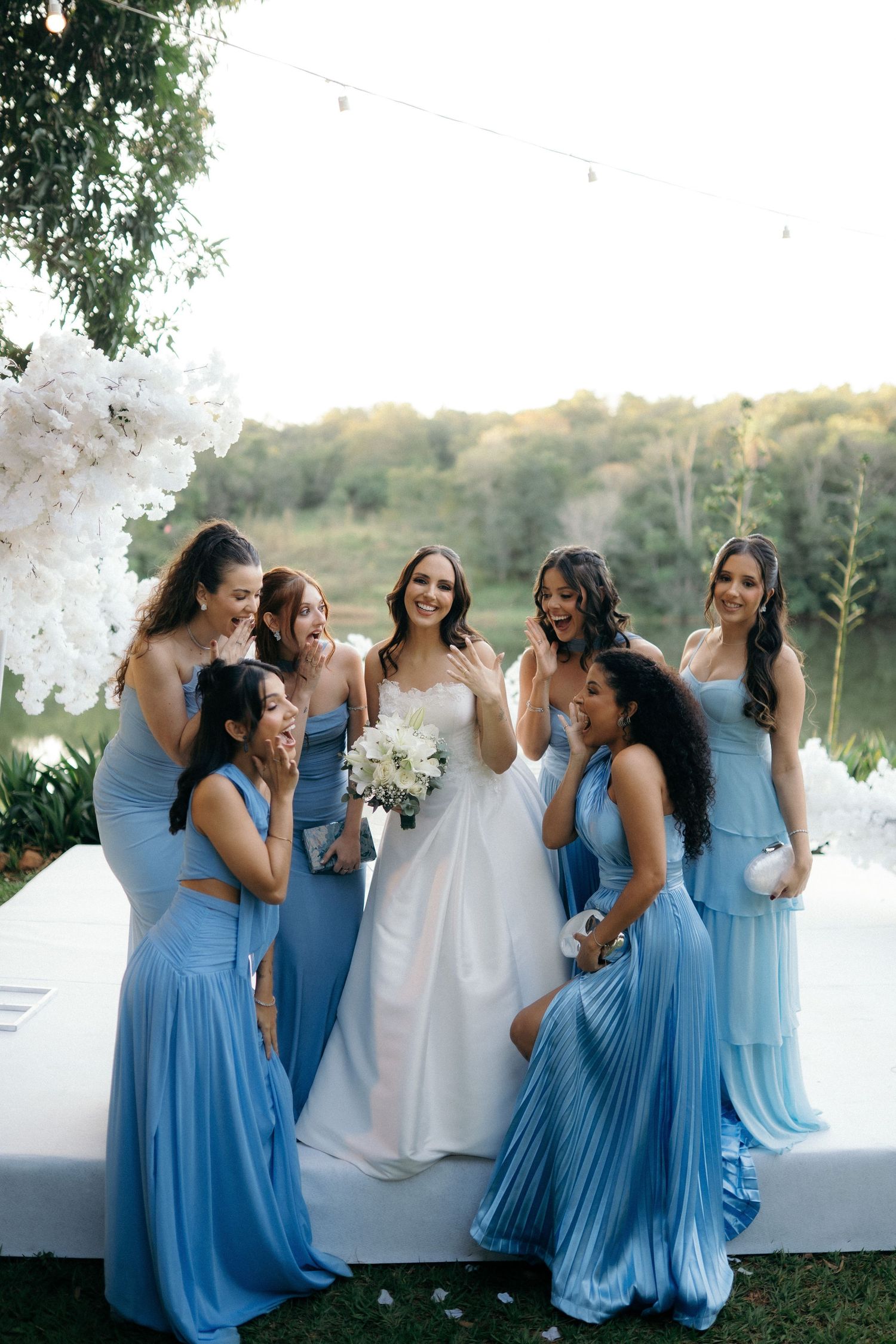 Bride in white gown laughing with six bridesmaids in blue dresses at outdoor wedding venue.