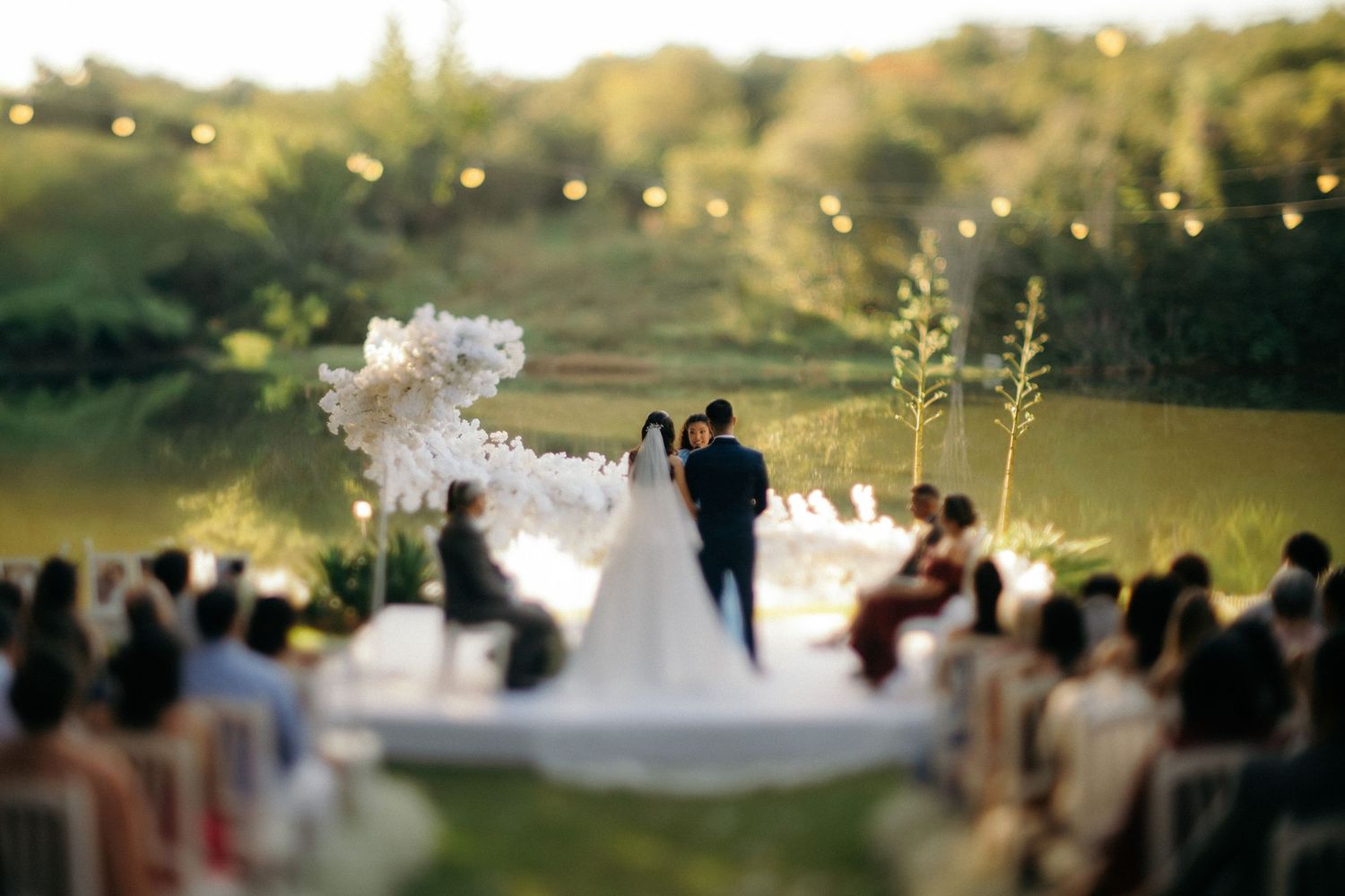 Outdoor wedding ceremony with bride and groom at altar by a lake, surrounded by guests and string lights at sunset.