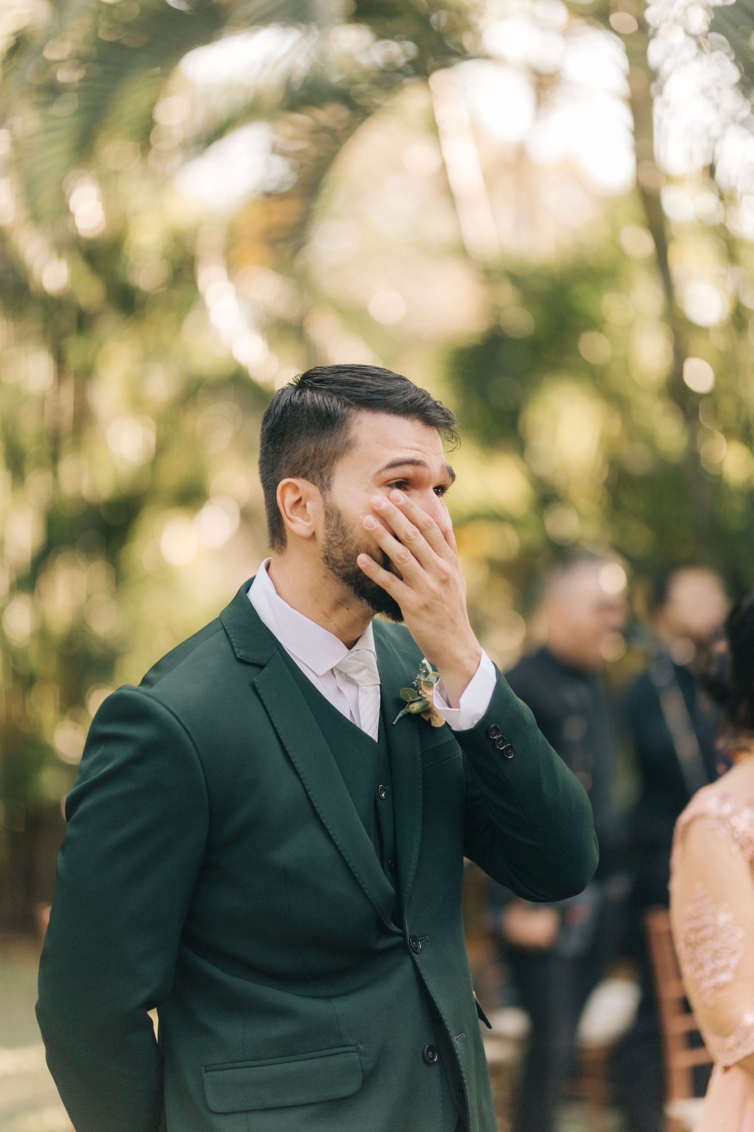 Emotional groom in green suit covering his mouth as bride walks down the aisle outdoors.