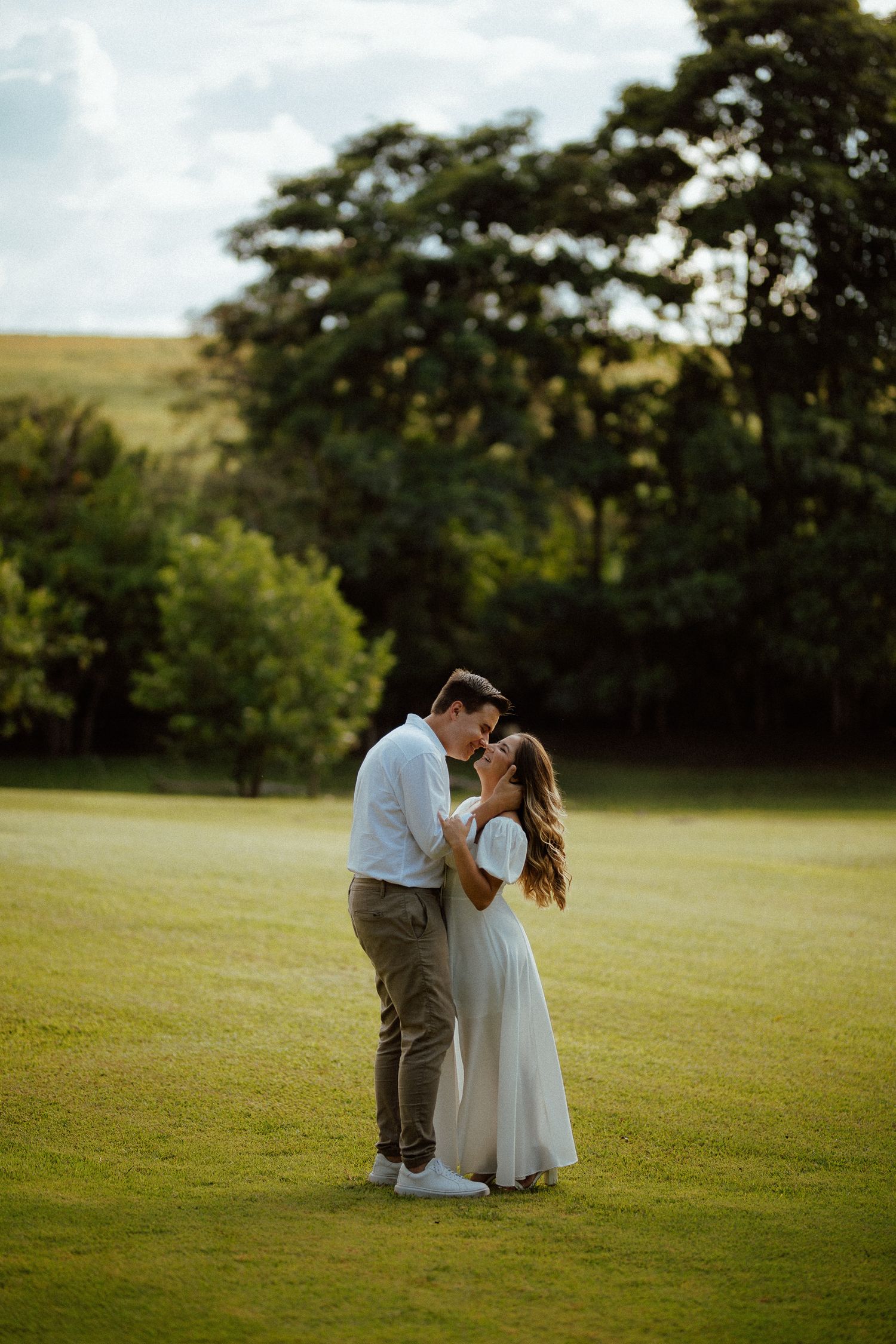 Romantic couple embracing forehead-to-forehead in golden hour sunlight on lush green open field.