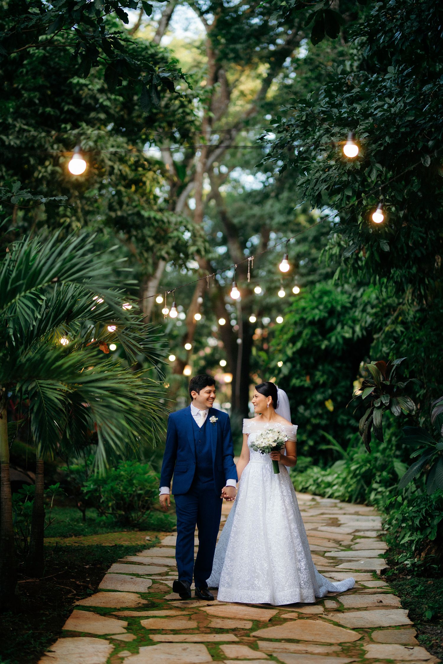 Newlyweds walking hand in hand along garden path with string lights and lush green trees.