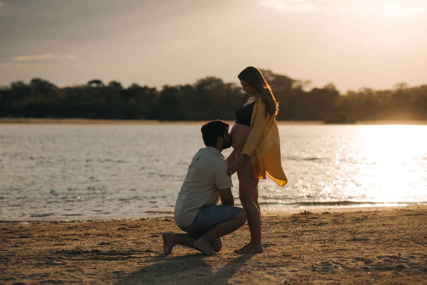 Man kneeling tenderly before a pregnant woman on a beach during a warm sunset.