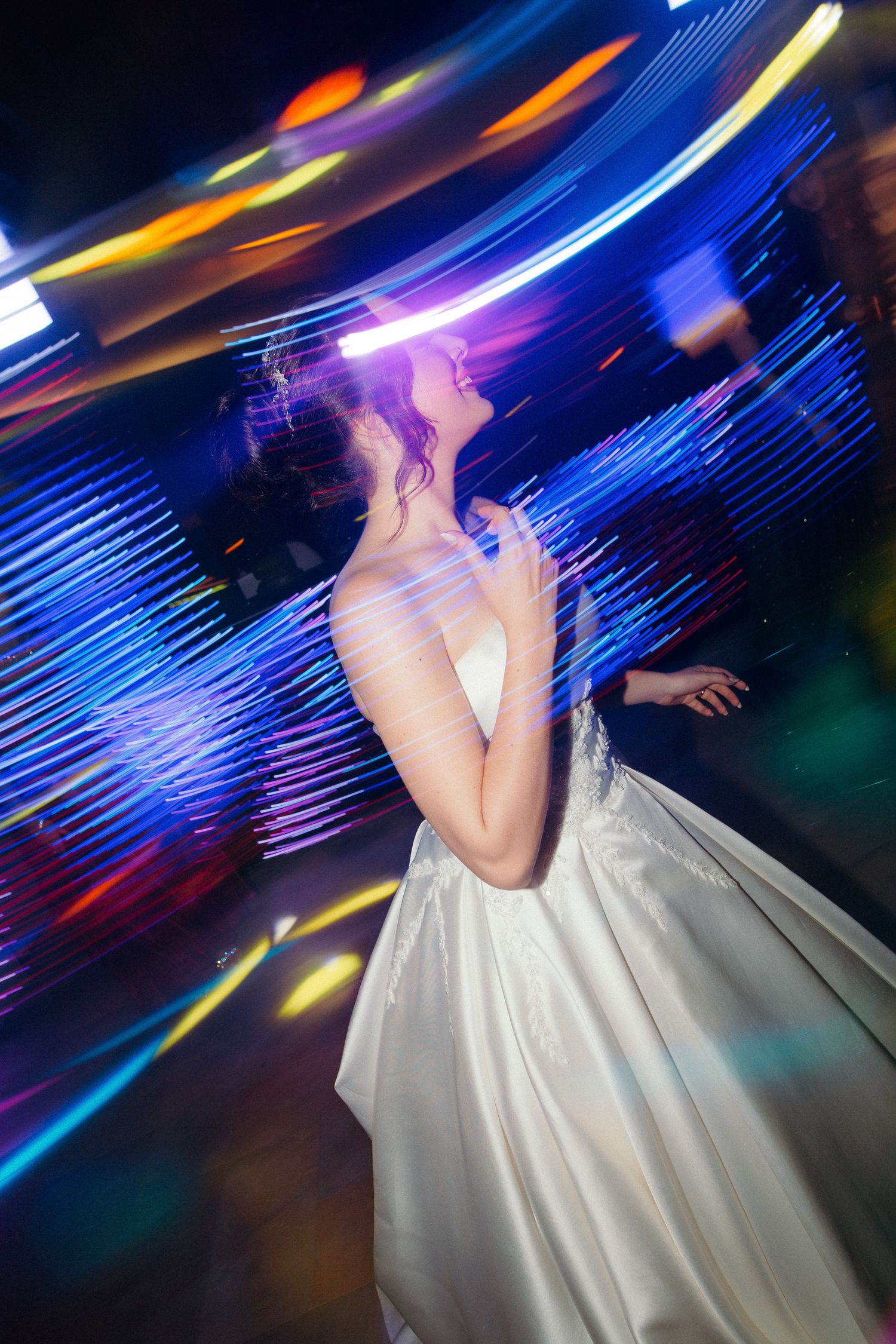 Bride dancing joyfully on the dance floor surrounded by colorful motion-blur lights at wedding reception.