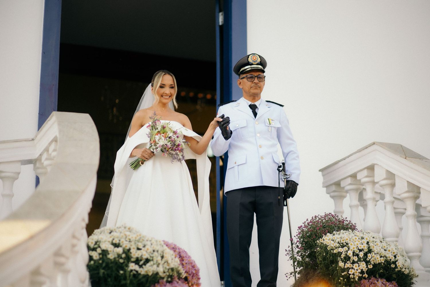 Bride in white ball gown with bouquet stands beside uniformed military officer at church entrance steps.