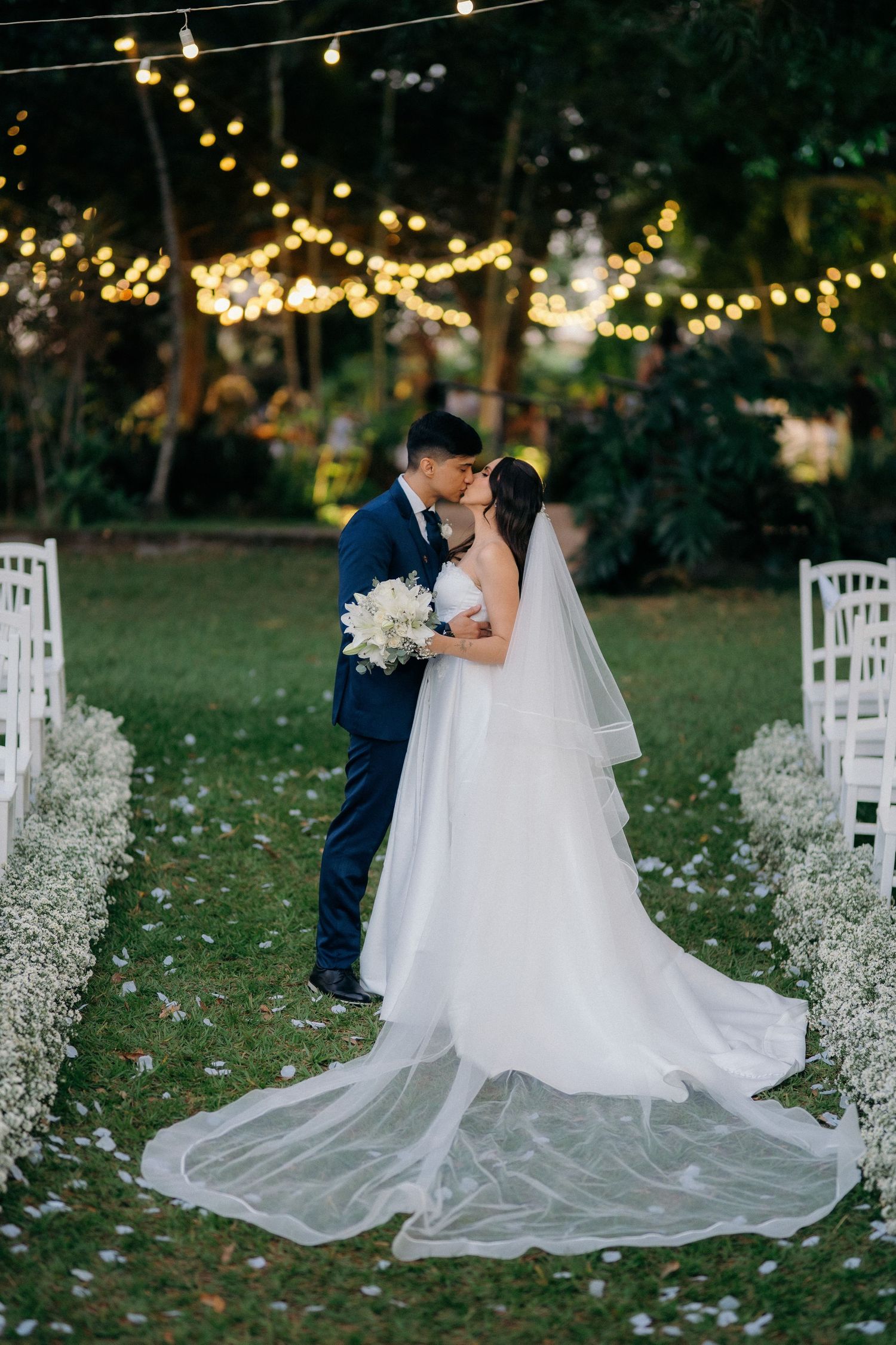 Newlyweds share a romantic kiss on a garden wedding aisle adorned with white flowers and string lights at dusk.