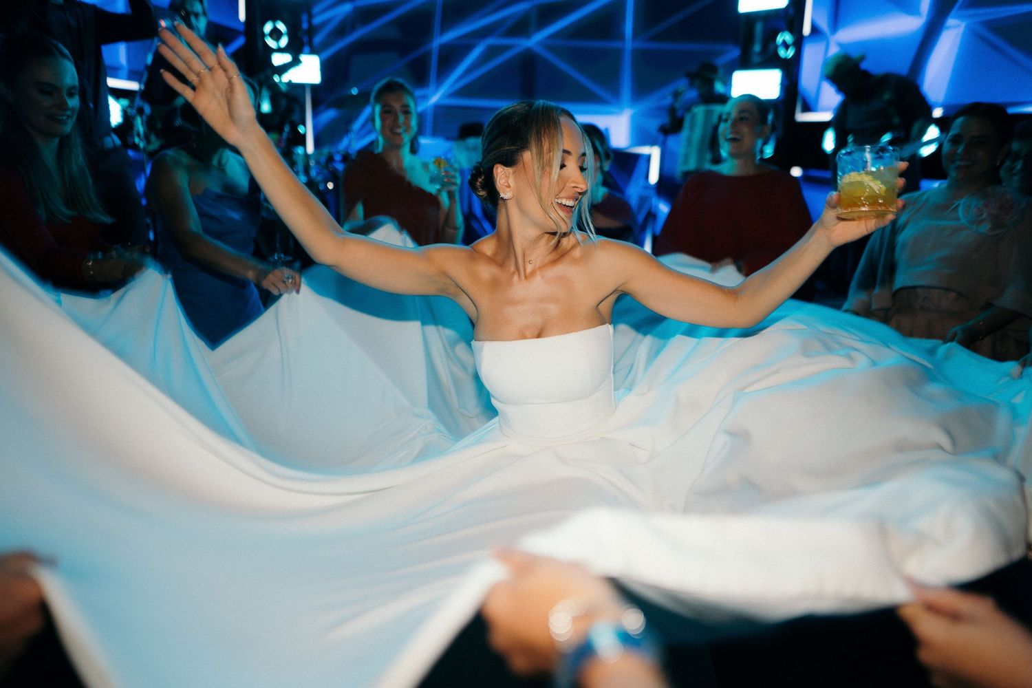Joyful bride in white strapless gown twirls on dance floor at vibrant blue-lit wedding reception.