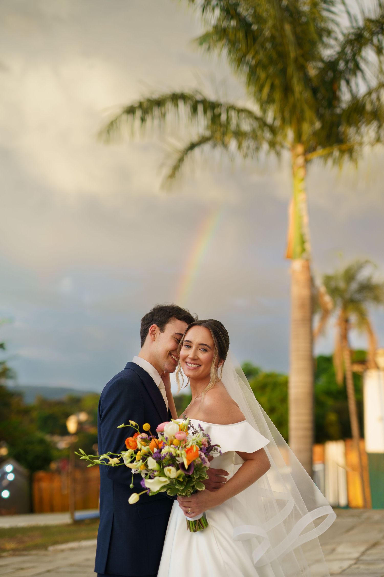 Bride and groom embracing outdoors with rainbow and palm tree in background on wedding day.