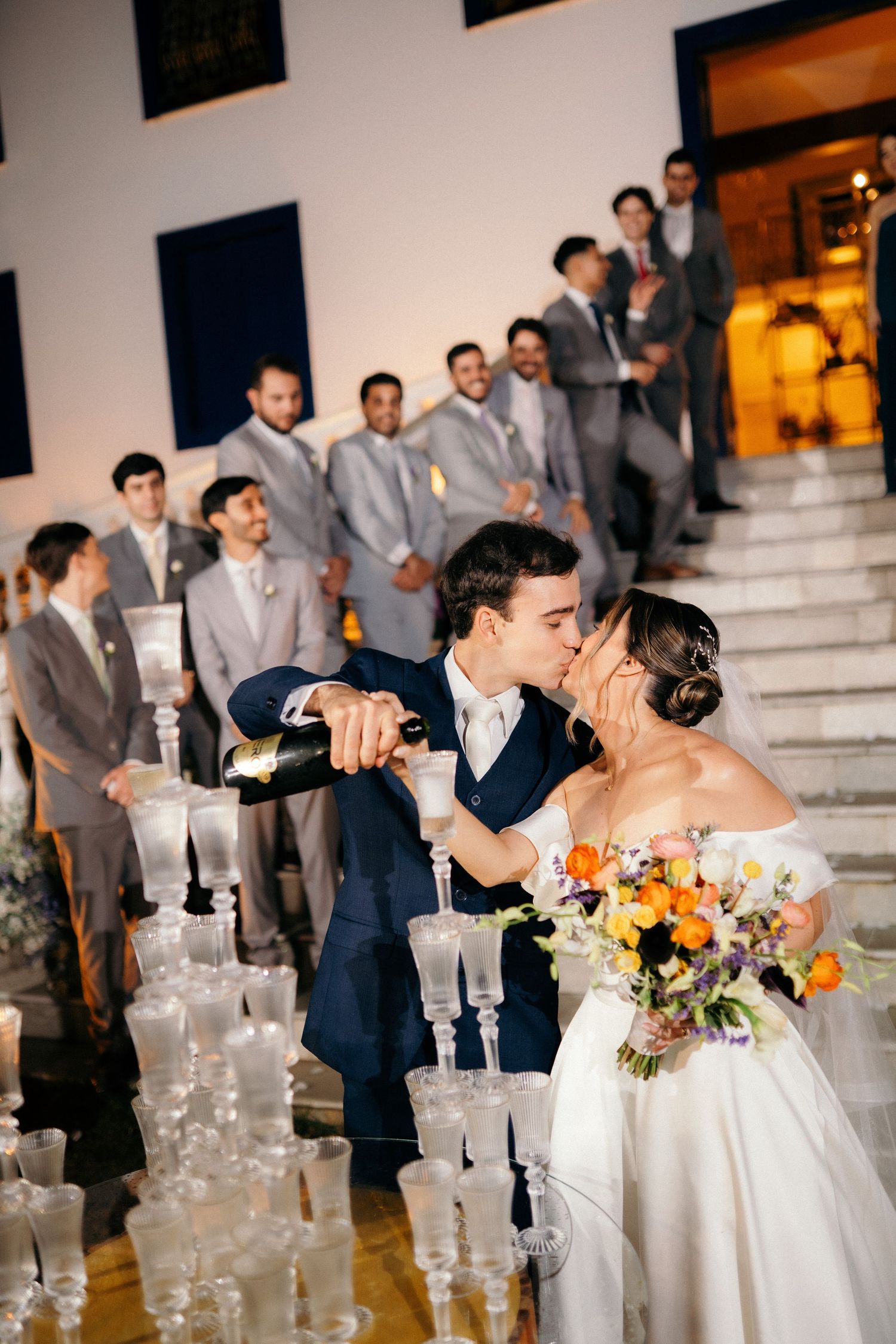 Bride and groom kissing while pouring champagne tower at wedding reception with groomsmen in gray suits on staircase.
