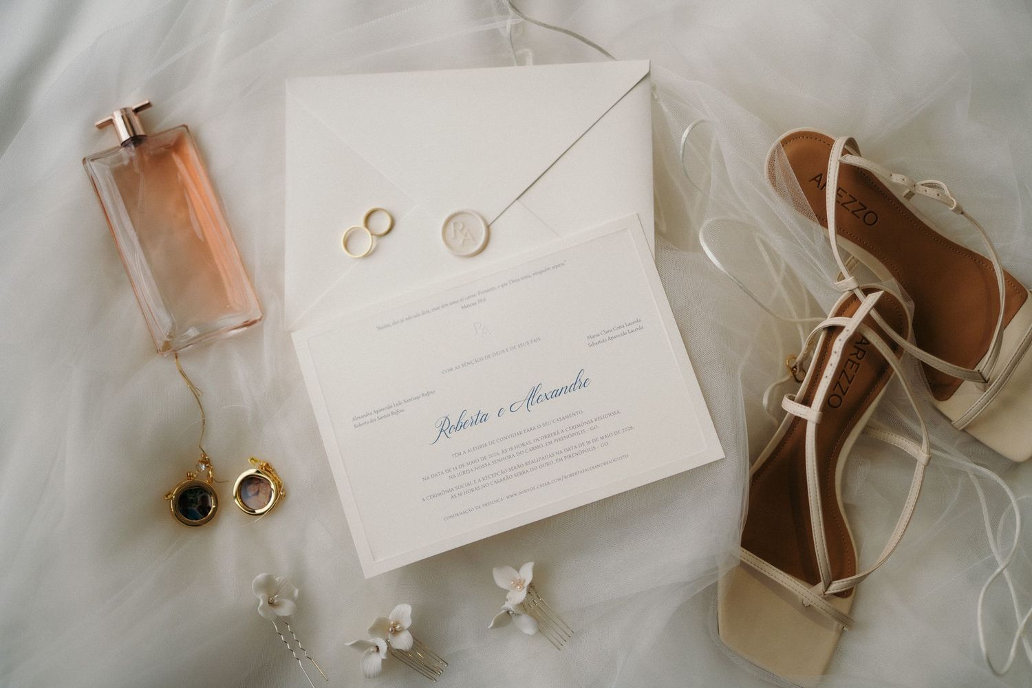 Elegant wedding flat lay with invitation, gold rings, perfume, earrings, and cream sandals on white fur.