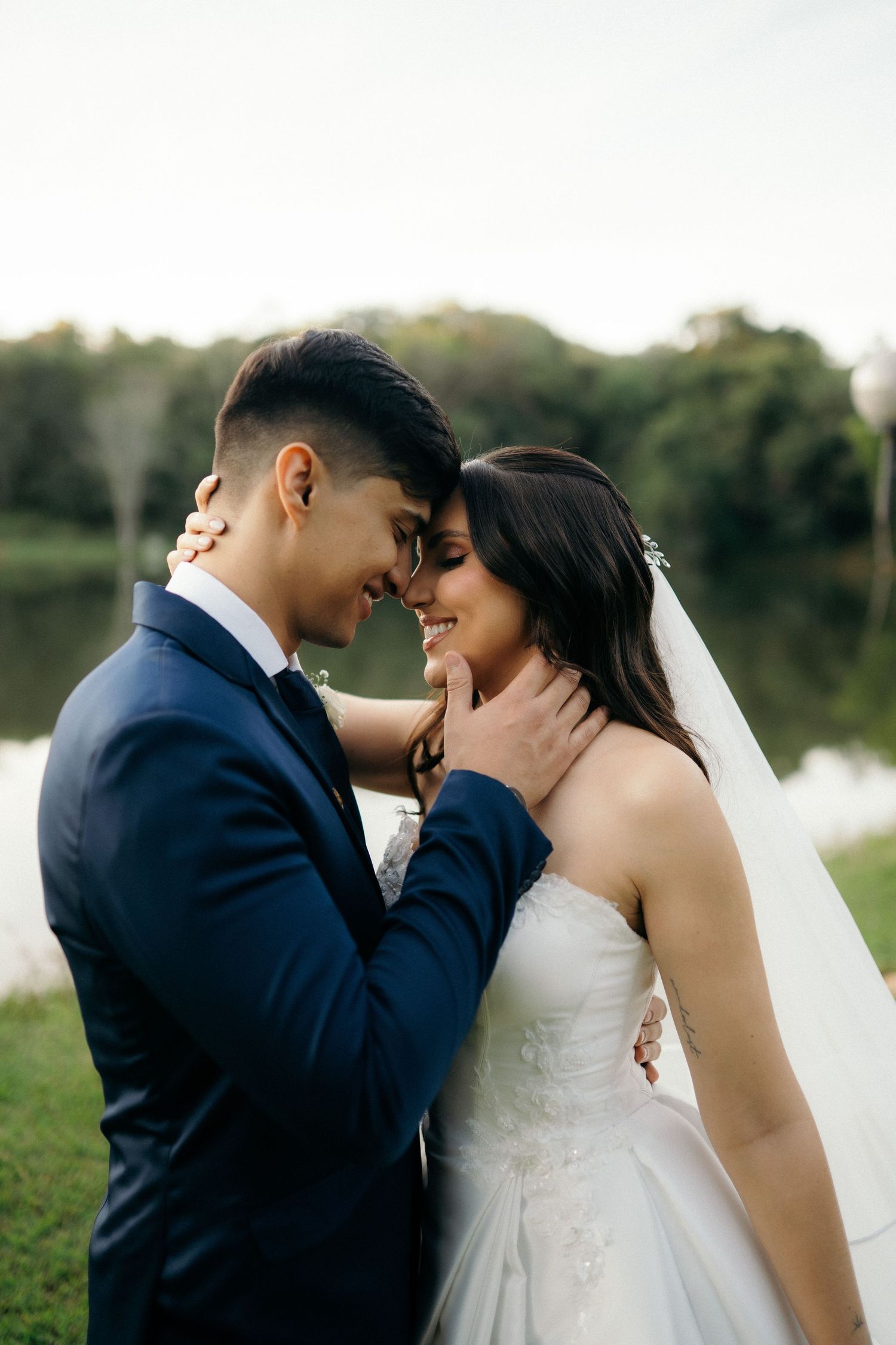 A bride and groom share a romantic forehead touch outdoors near a lake, she in a white gown, he in a navy suit.