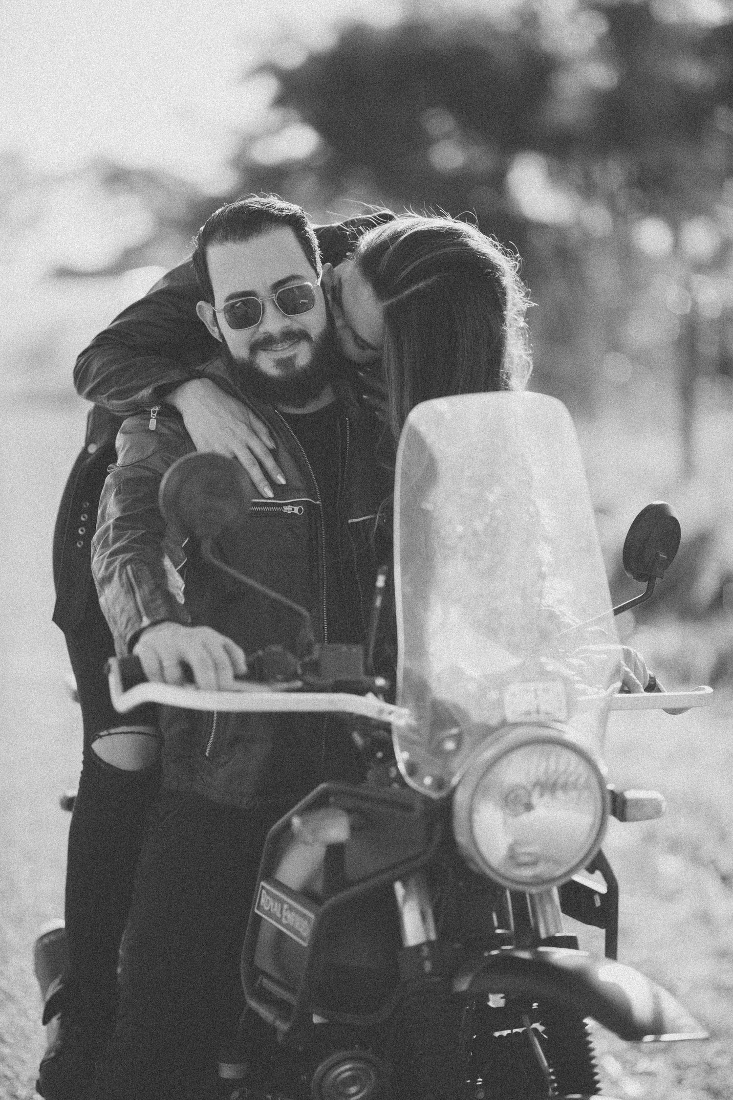 Black and white photo of couple embracing on a vintage motorcycle outdoors.