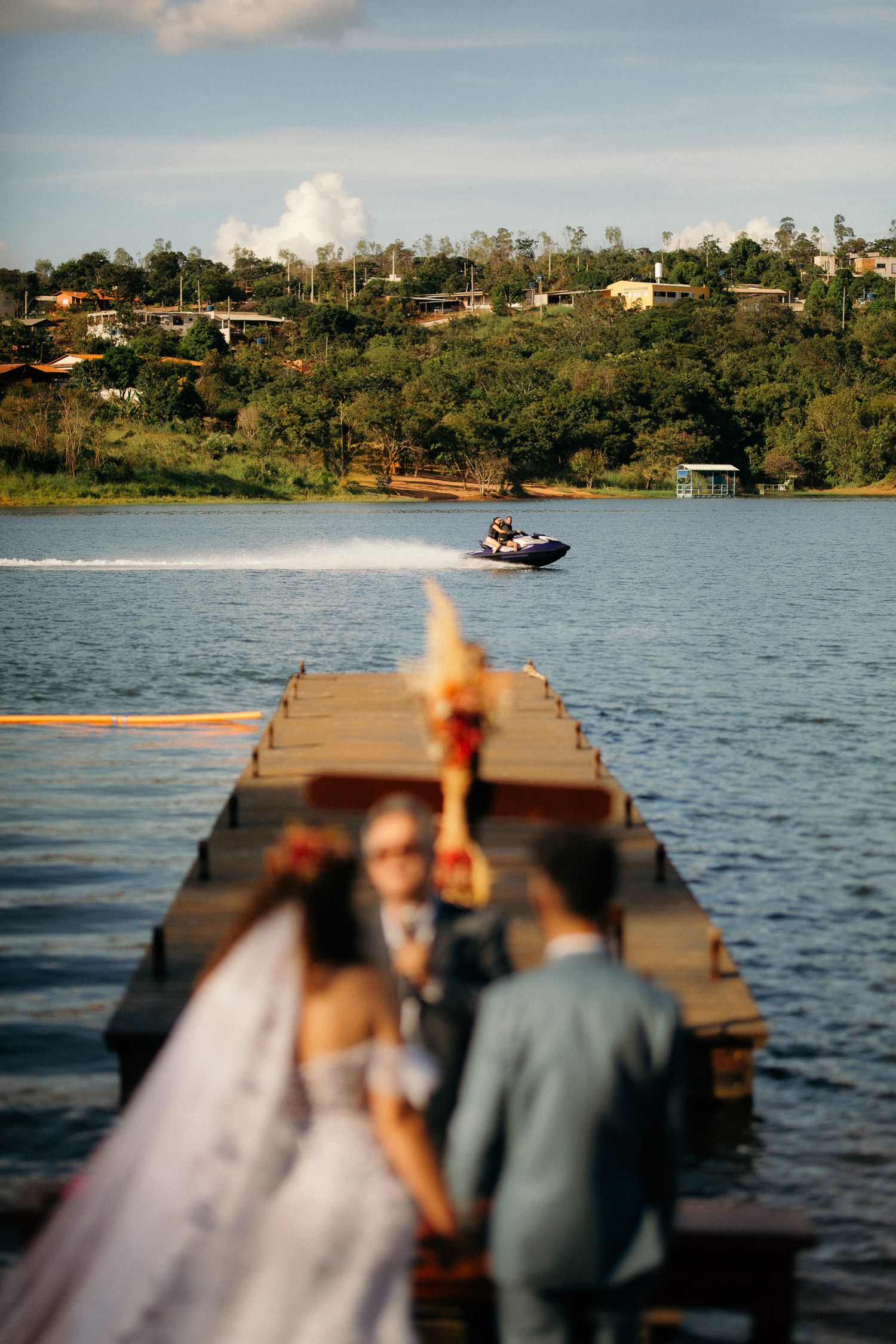 Bride and groom standing on a wooden pier over a lake with a jet ski in background.