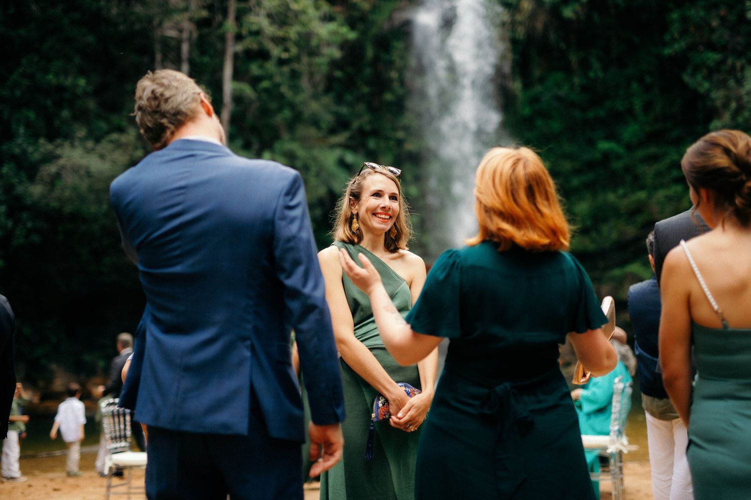 Wedding guests in green attire conversing near stunning waterfall outdoor ceremony venue.