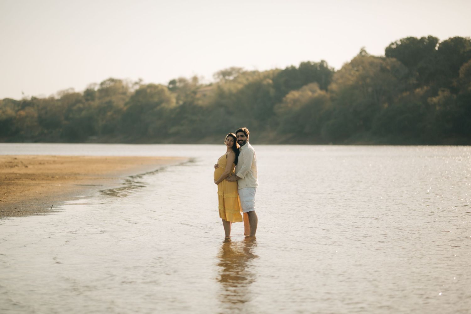 Romantic couple embracing while wading in shallow water along a scenic riverbank at sunset.