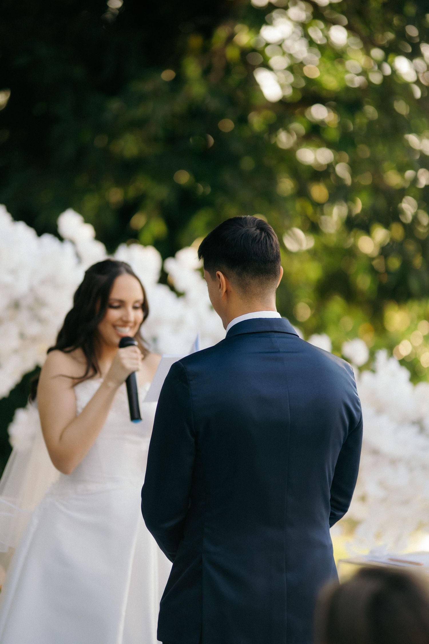 Bride smiling and holding microphone reciting vows to groom at outdoor wedding ceremony with white floral arch.