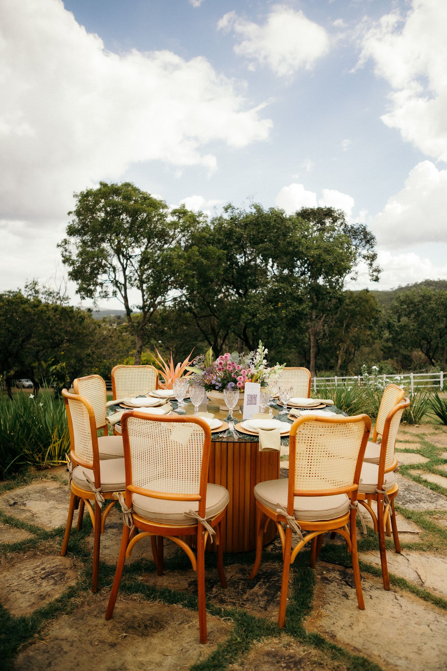 Elegant outdoor wedding reception table with rattan chairs, floral centerpiece, and lush green trees under a cloudy sky.