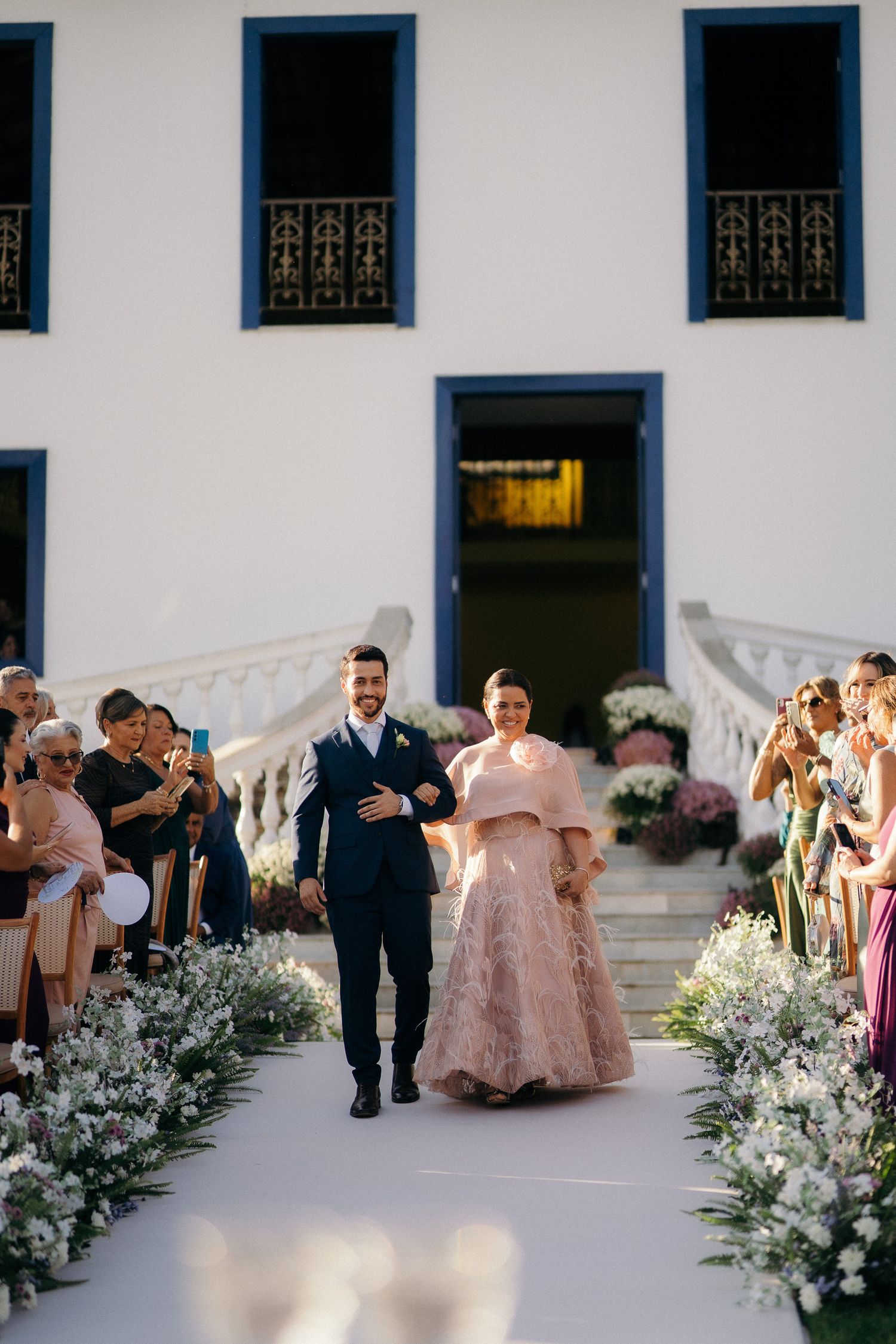 Couple walking down outdoor wedding aisle, bride in pink lace gown at elegant white venue.