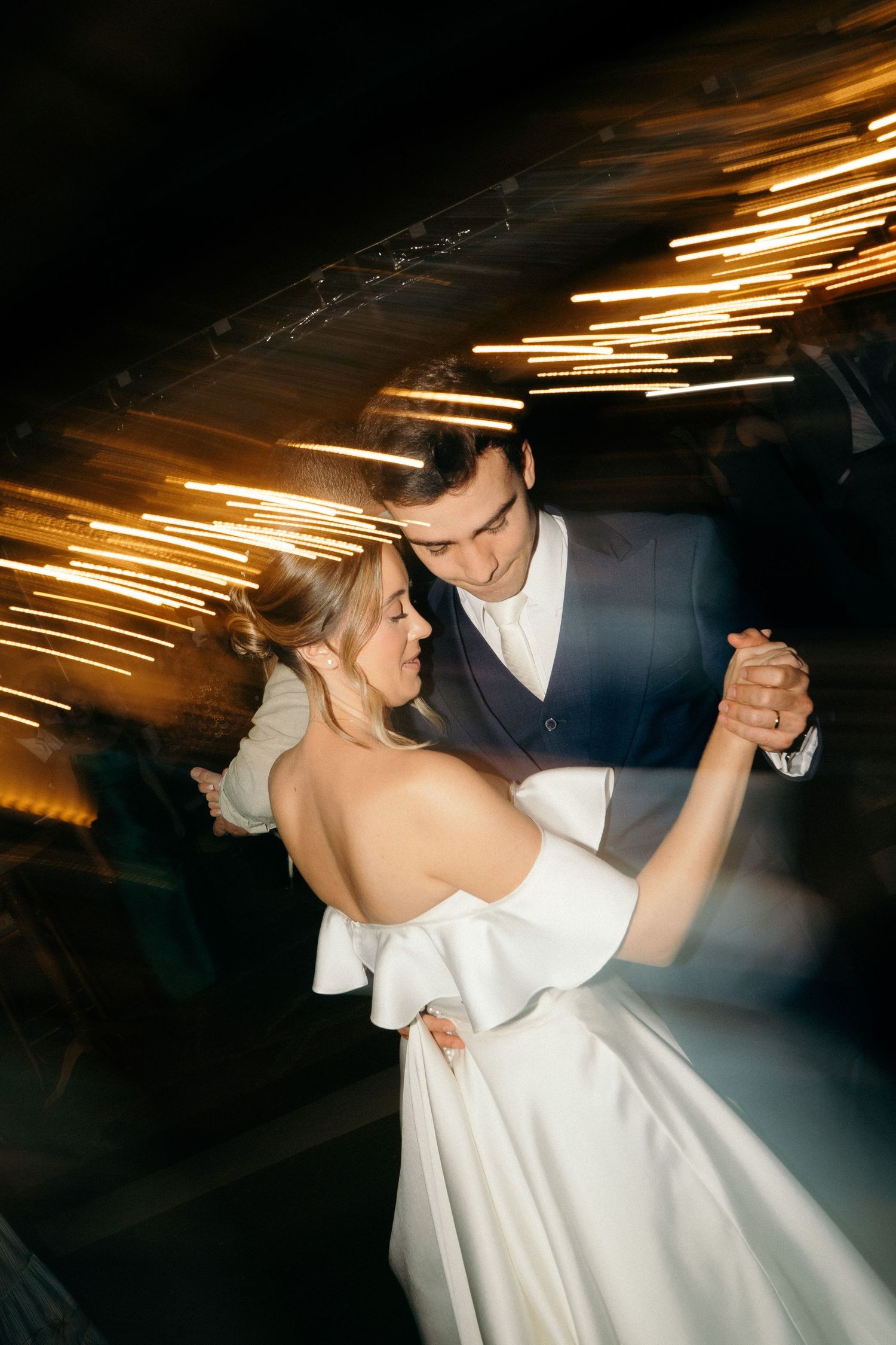 Bride and groom share first dance with golden light streaks in dark reception venue.