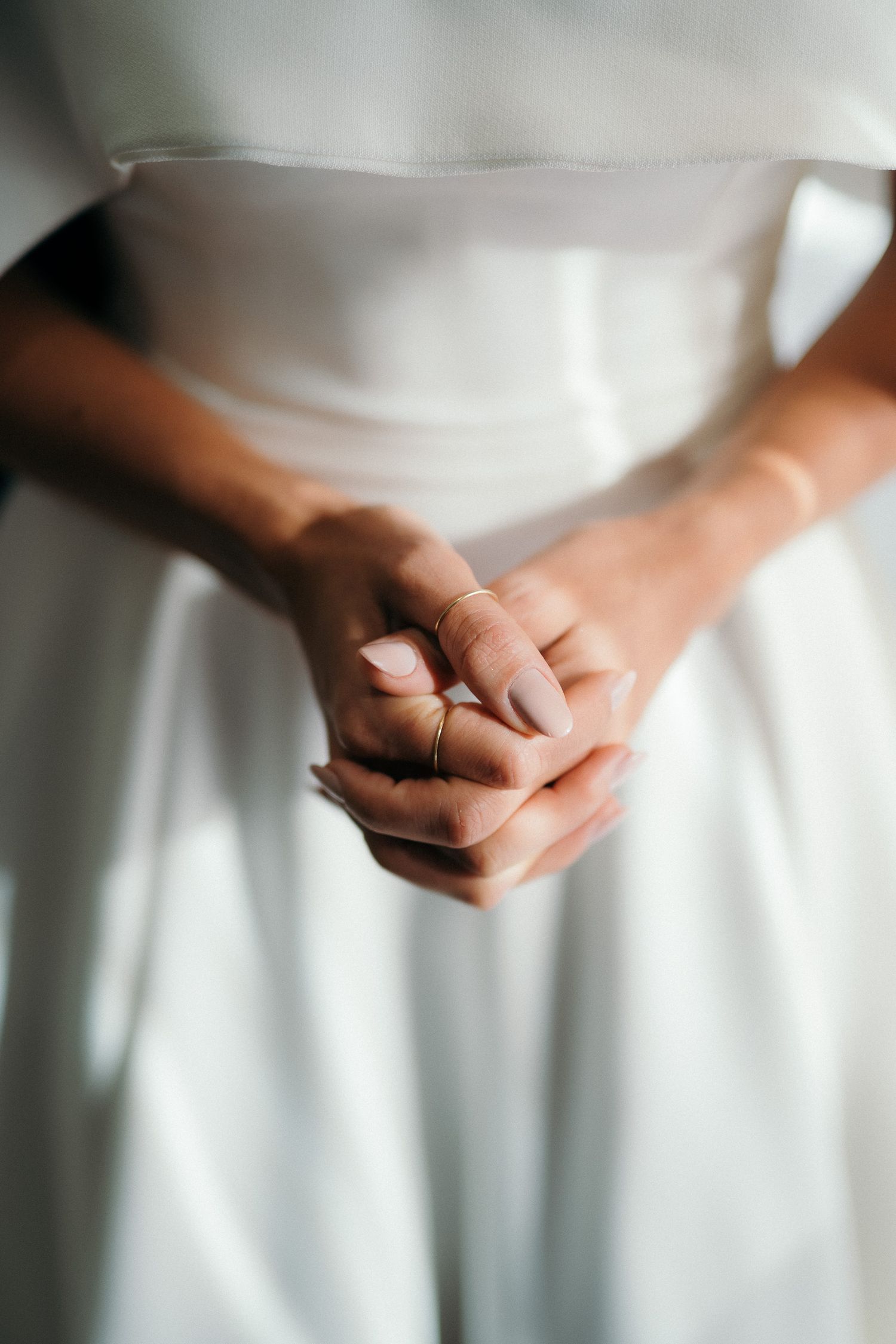 Close-up of bride's hands clasped together wearing gold rings over a white wedding gown.