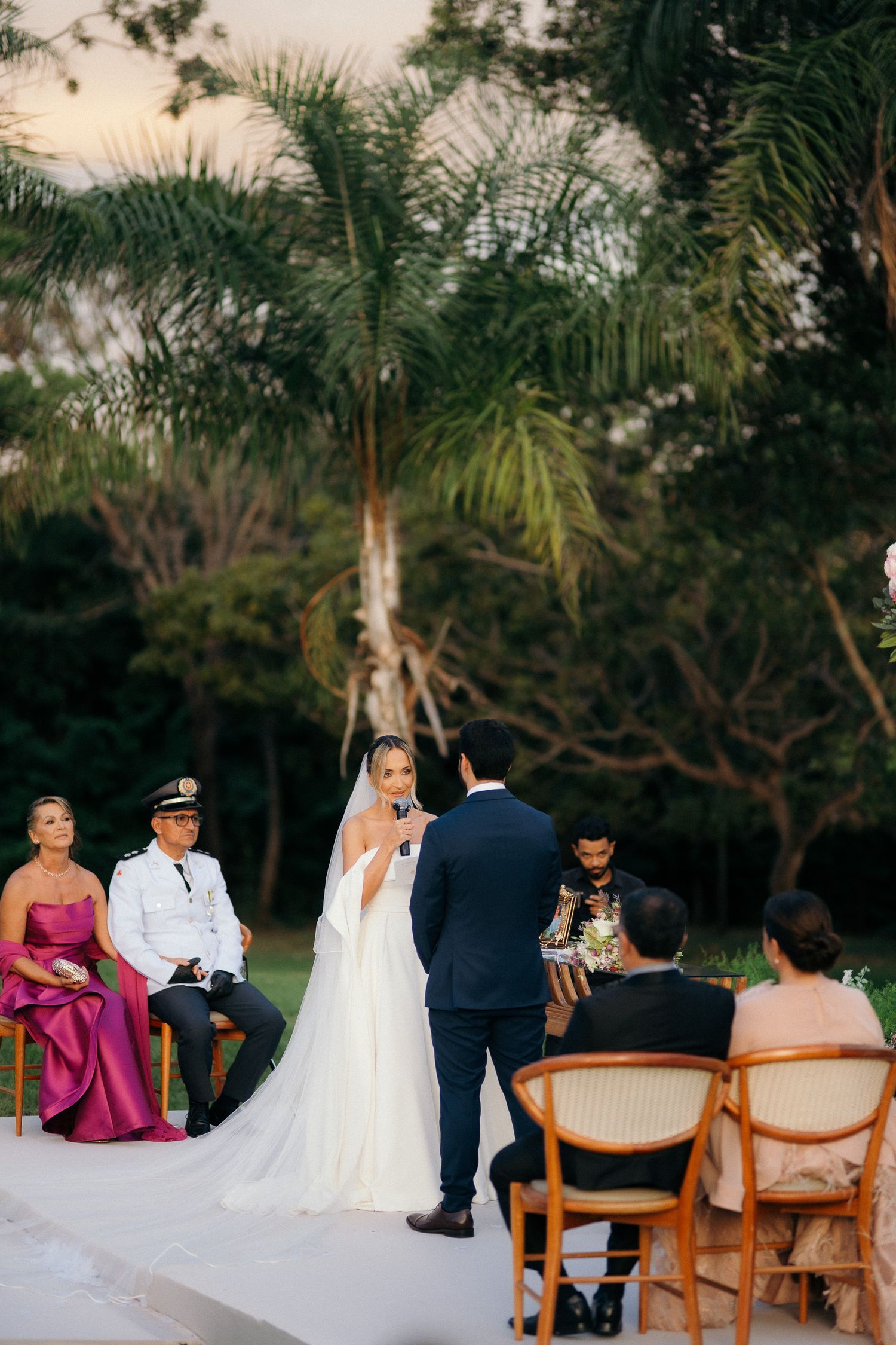 Bride in white gown reciting vows to groom at an outdoor evening wedding ceremony with palm trees.