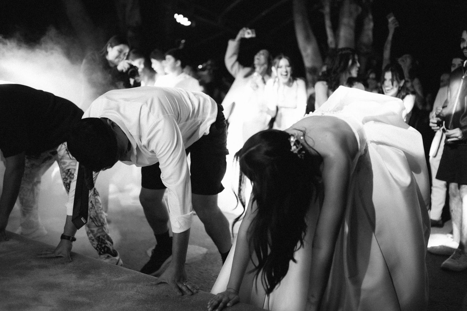 Black and white photo of wedding guests dancing energetically in smoke-filled atmosphere at reception.