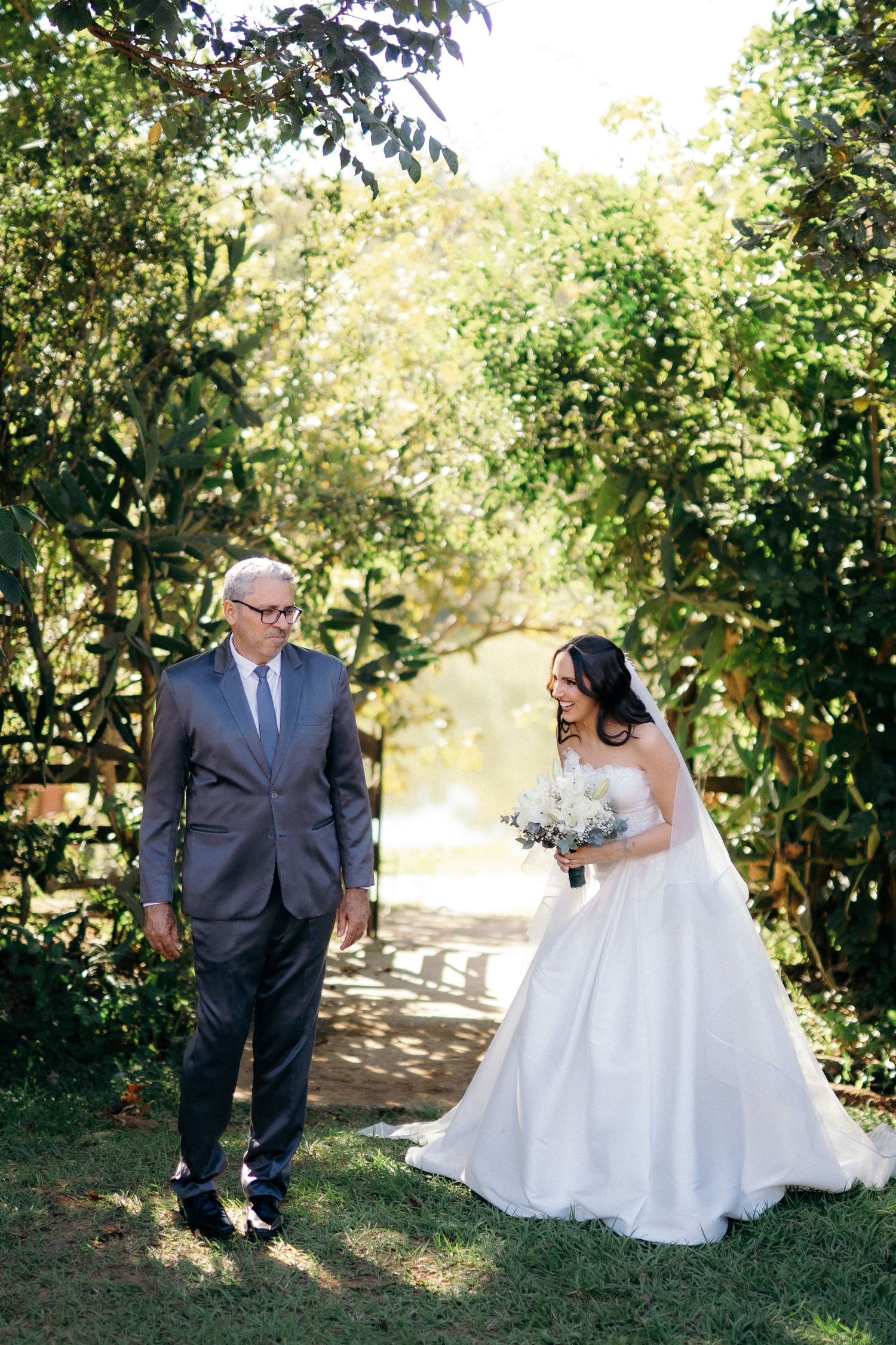 Bride in white gown holding bouquet meets older man in gray suit along a lush green garden path.