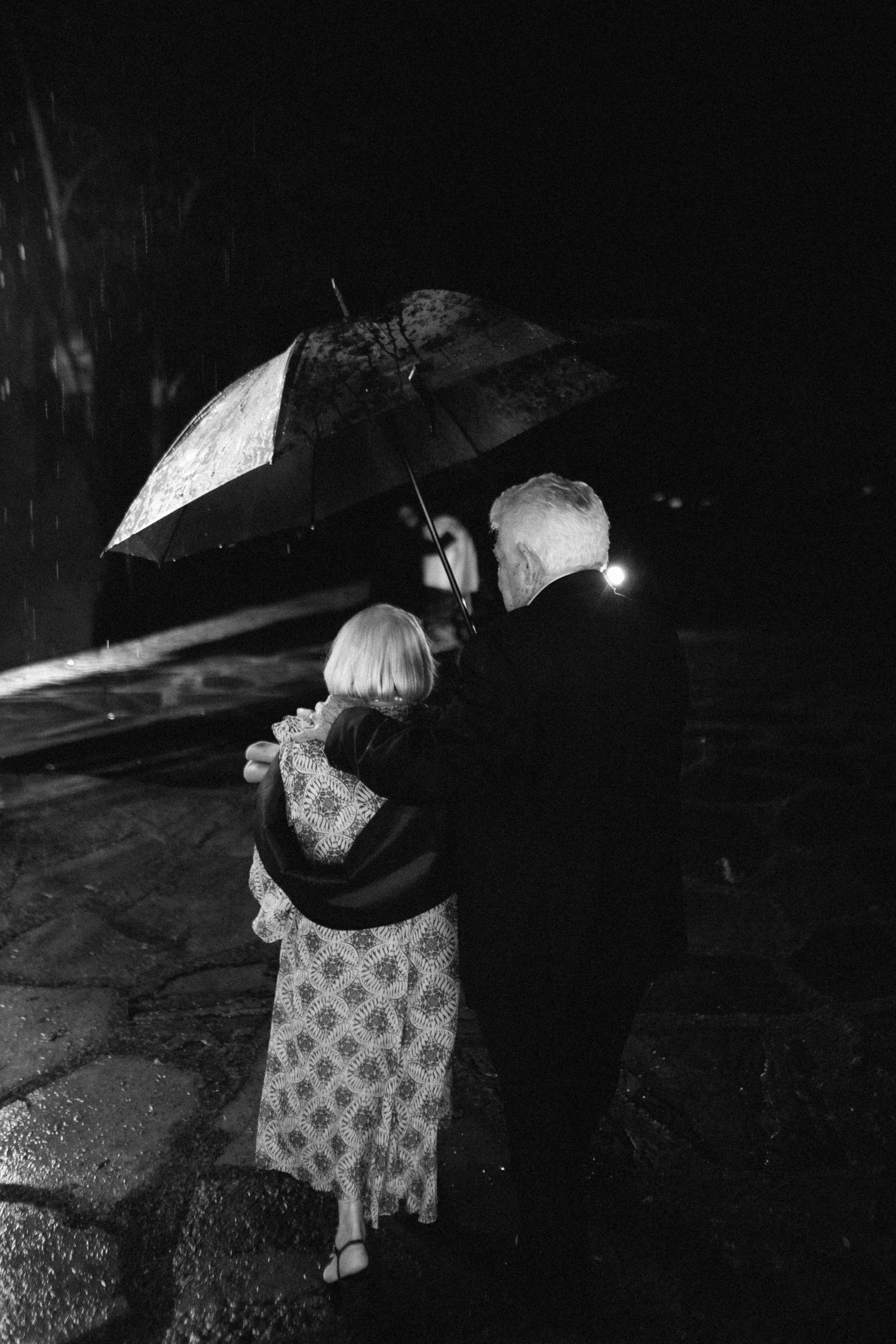Black and white photo of an elderly couple sharing an umbrella in the rain at a nighttime wedding event.