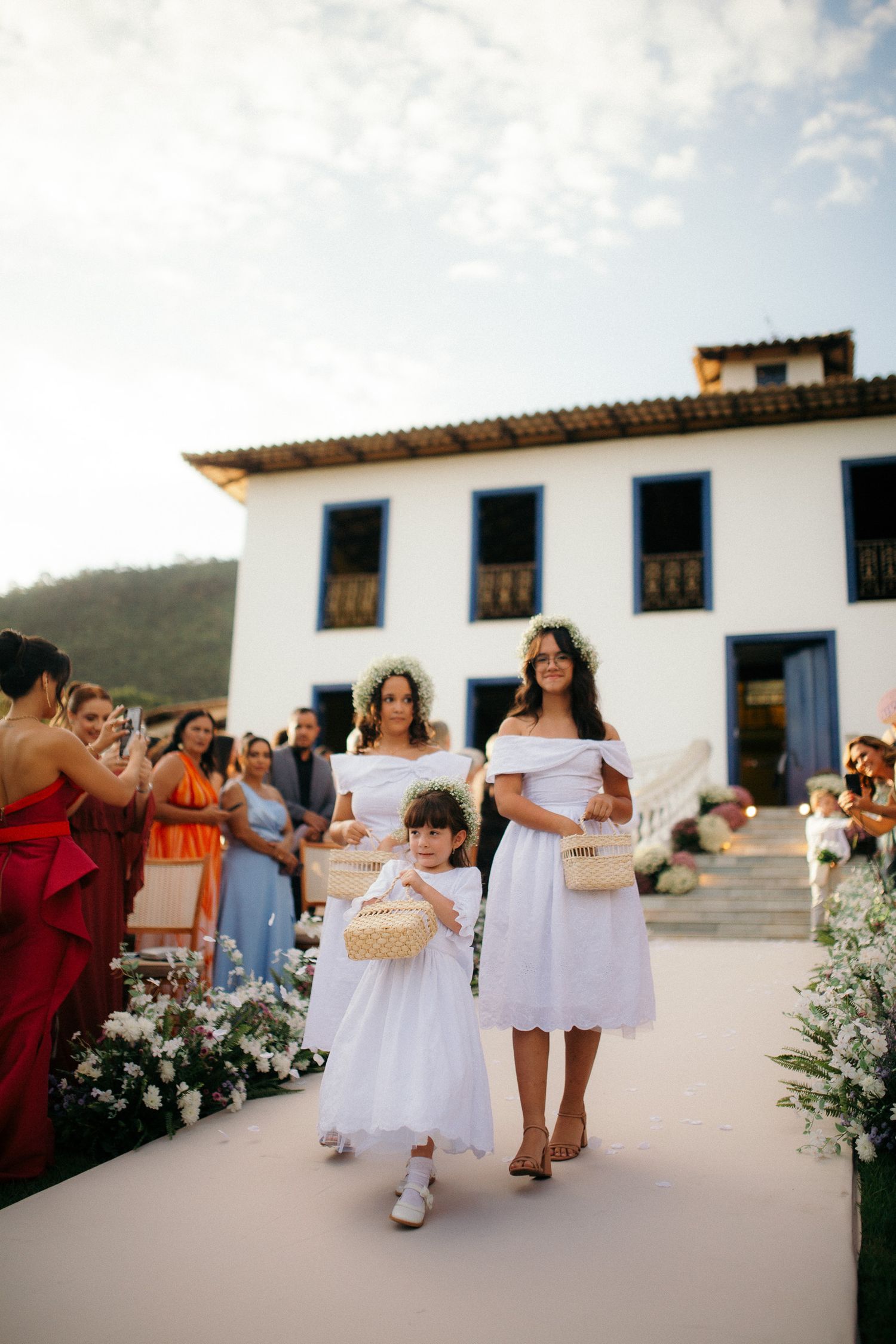 Two flower girls in white dresses walk down the aisle at an outdoor wedding ceremony at a colonial estate.