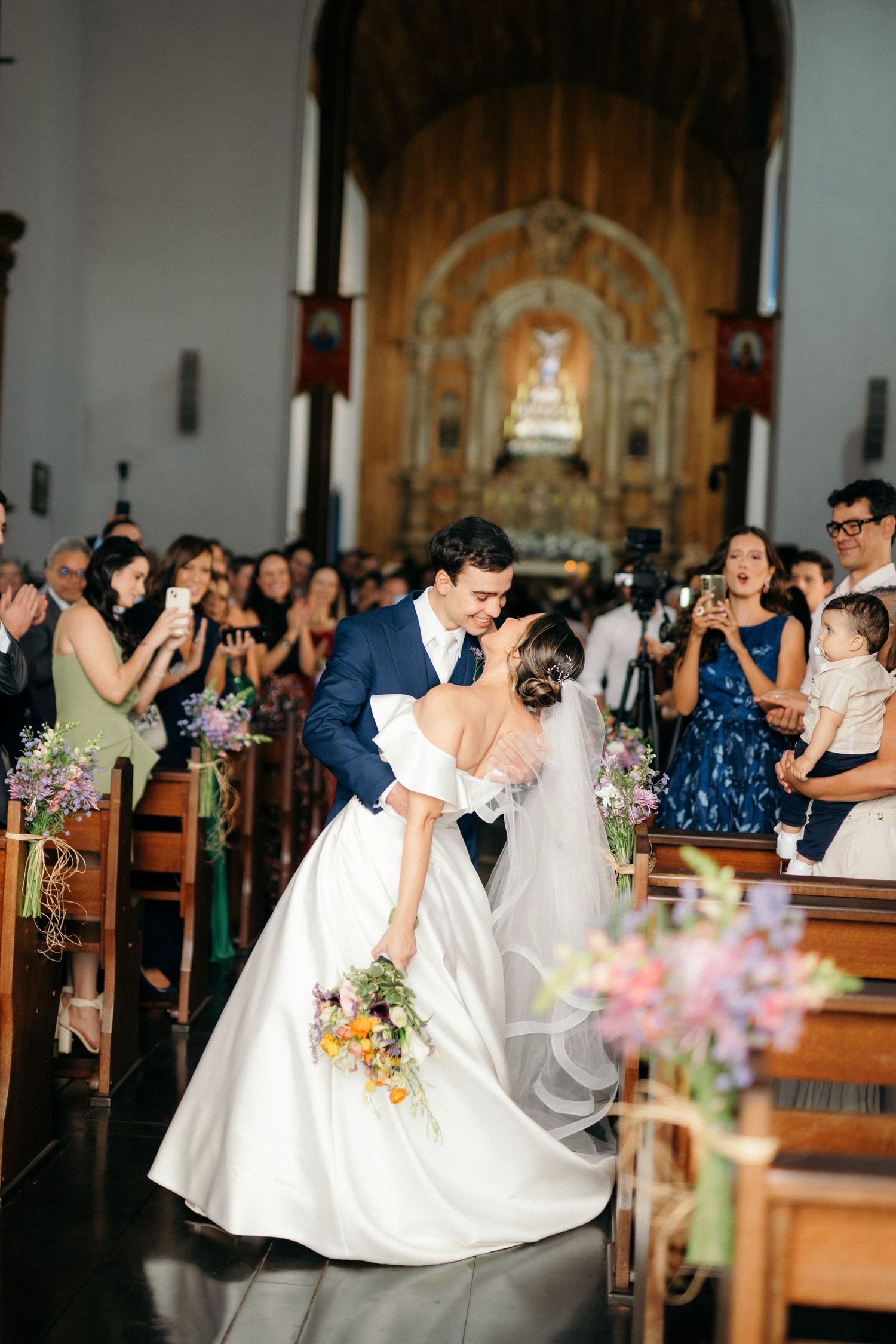 Groom dipping bride for a kiss in church aisle surrounded by cheering wedding guests.