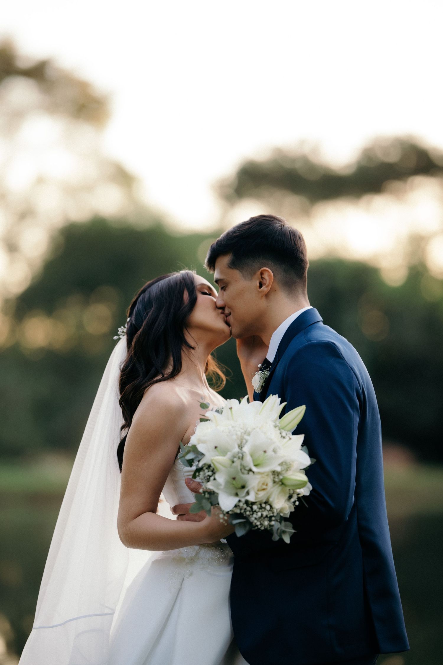 Bride and groom sharing a romantic kiss at golden hour holding white lily wedding bouquet.