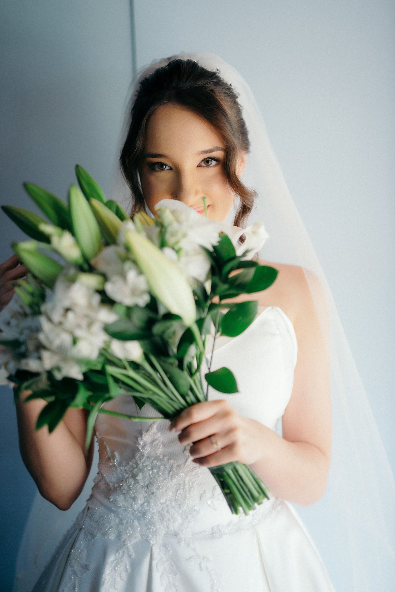 Bride in white lace gown and veil holding elegant bouquet of white lilies and flowers, partially covering her face.