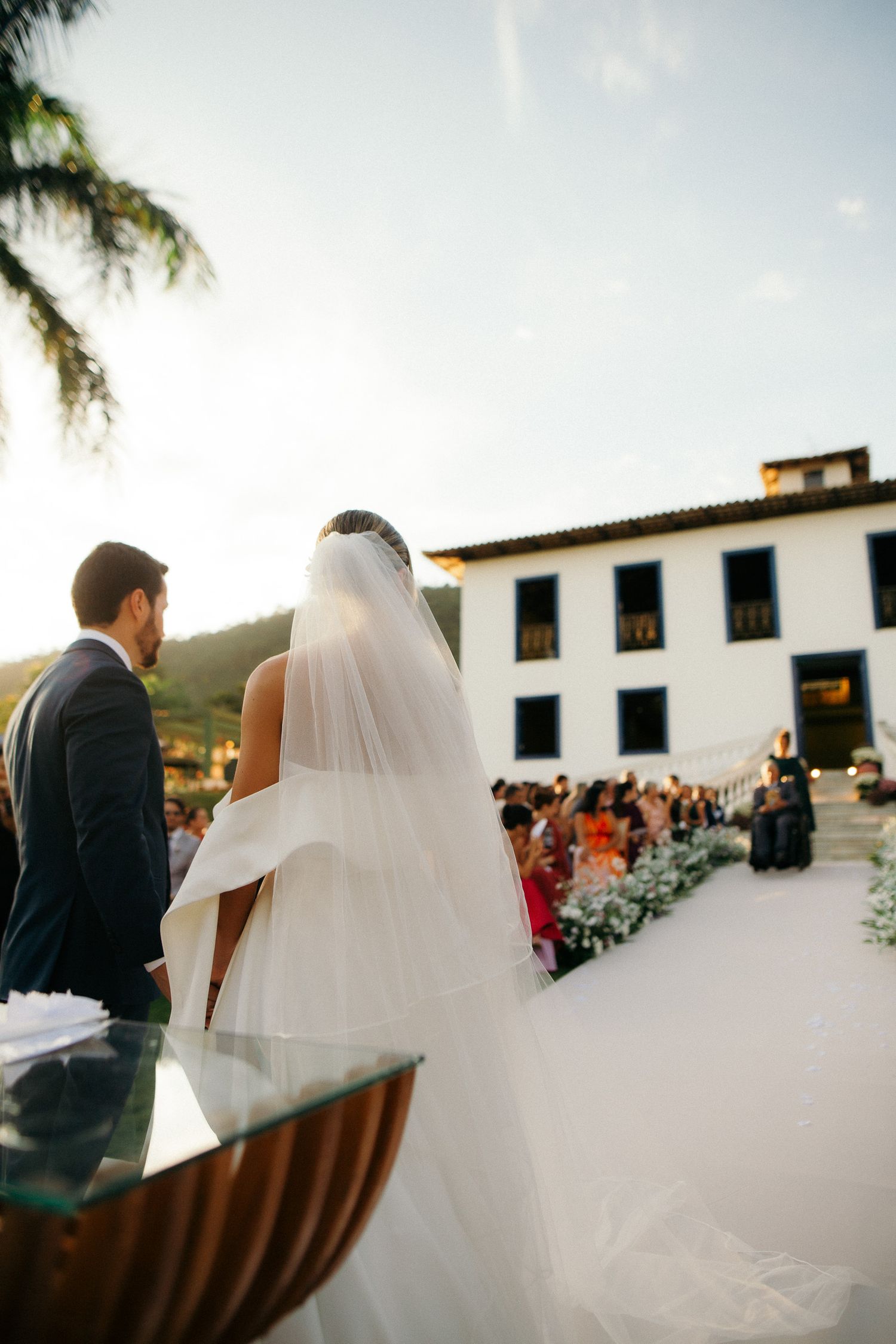 Bride and groom standing at outdoor wedding altar with guests seated and historic venue behind them.
