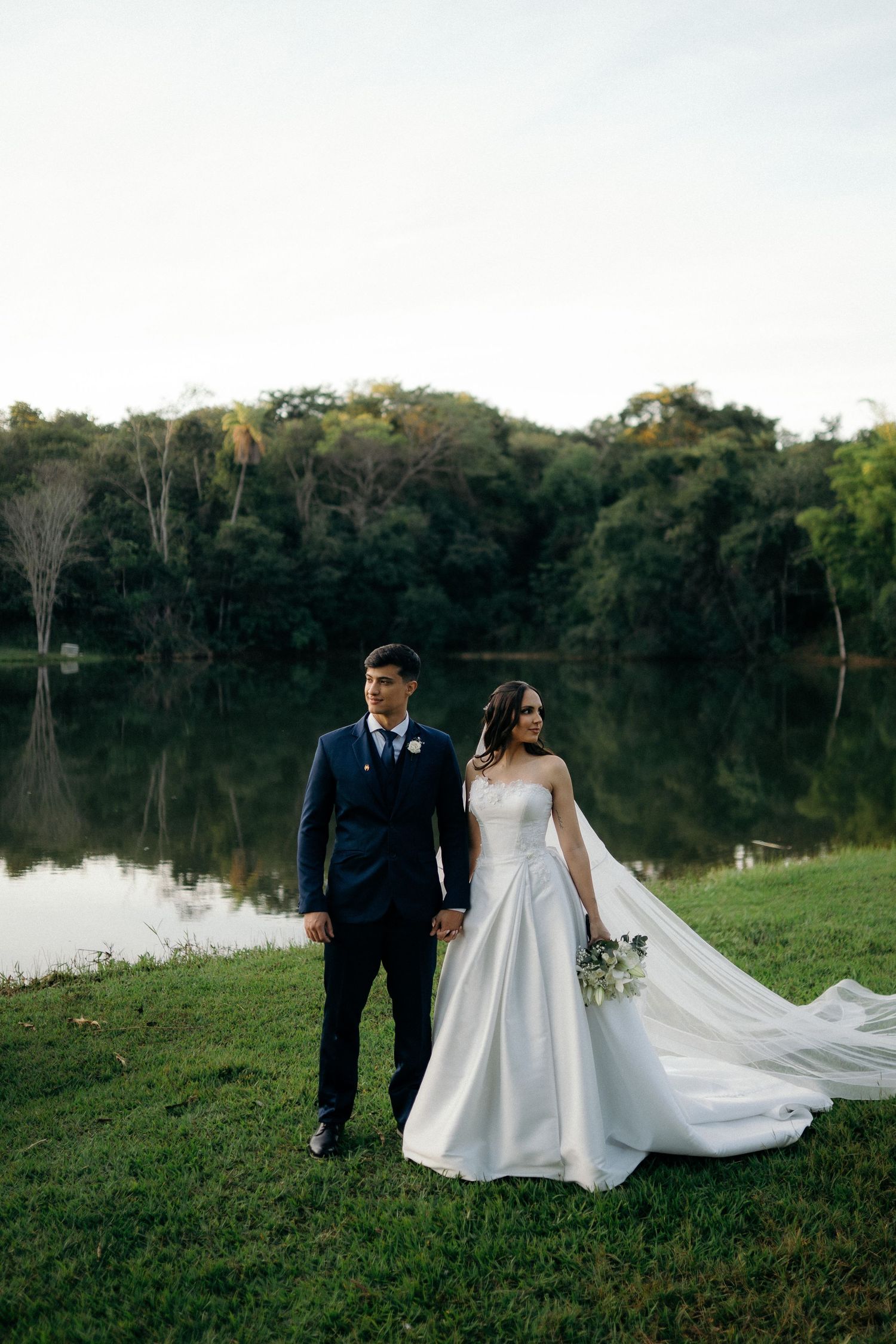Bride and groom hold hands by a tranquil lake at dusk, her gown train flowing across the green grass.