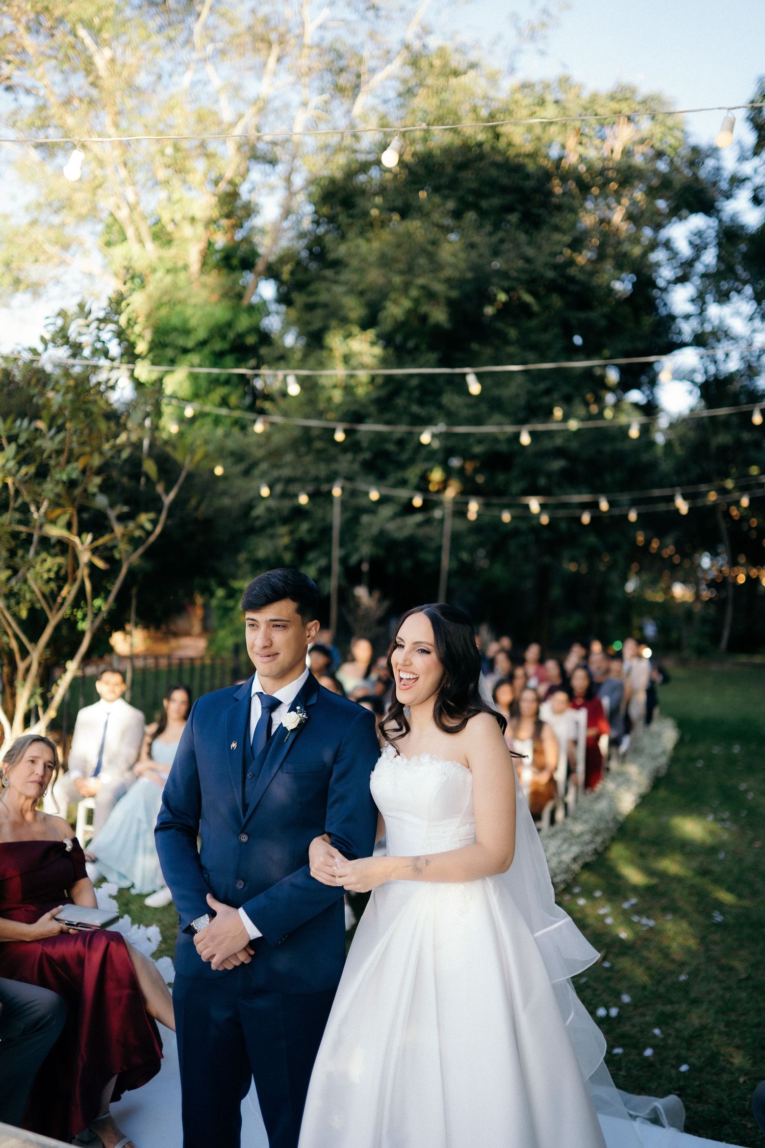 Bride and groom walking down aisle at outdoor evening wedding with string lights overhead.