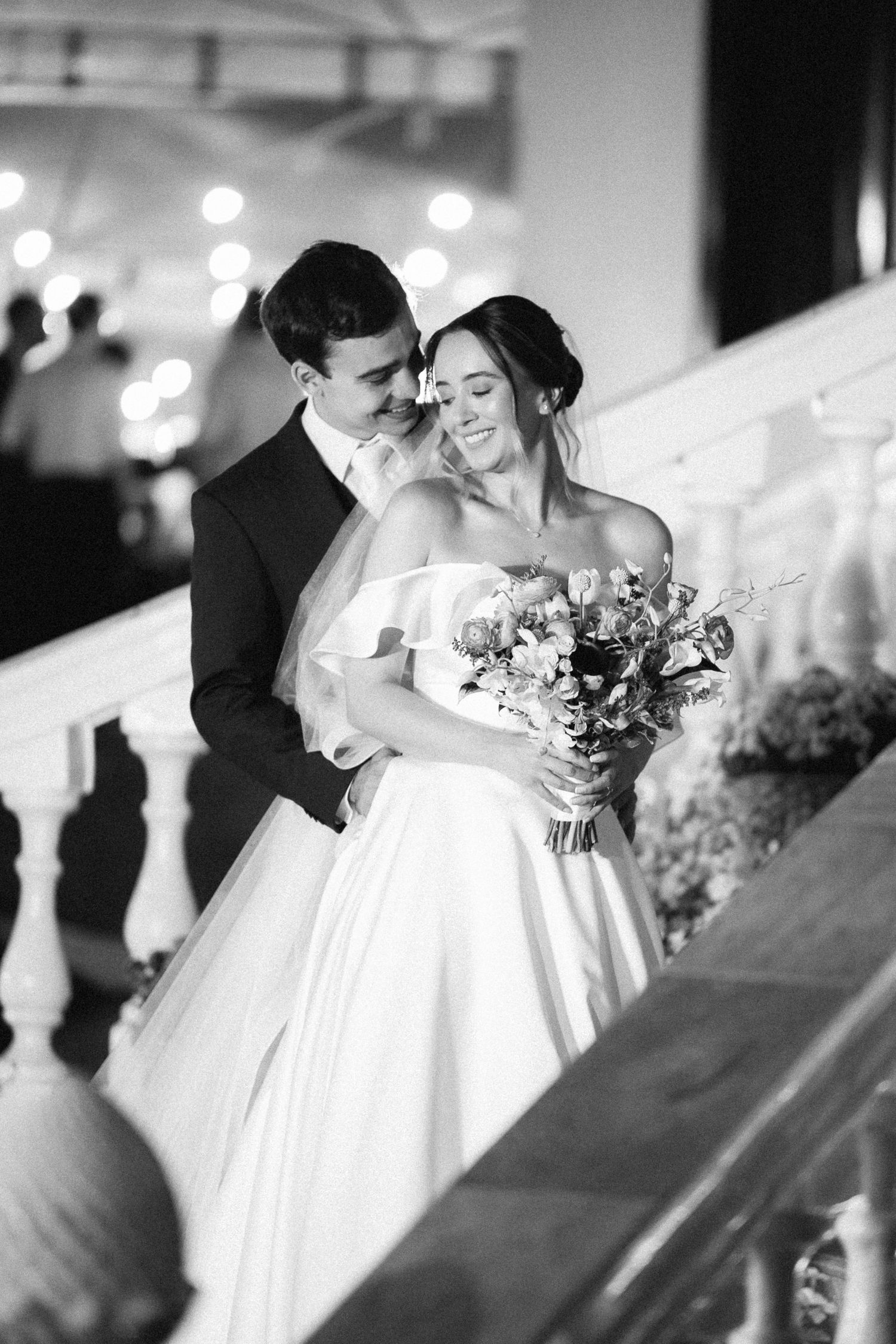 Black and white wedding photo of bride and groom embracing on elegant staircase, bride holding bouquet.